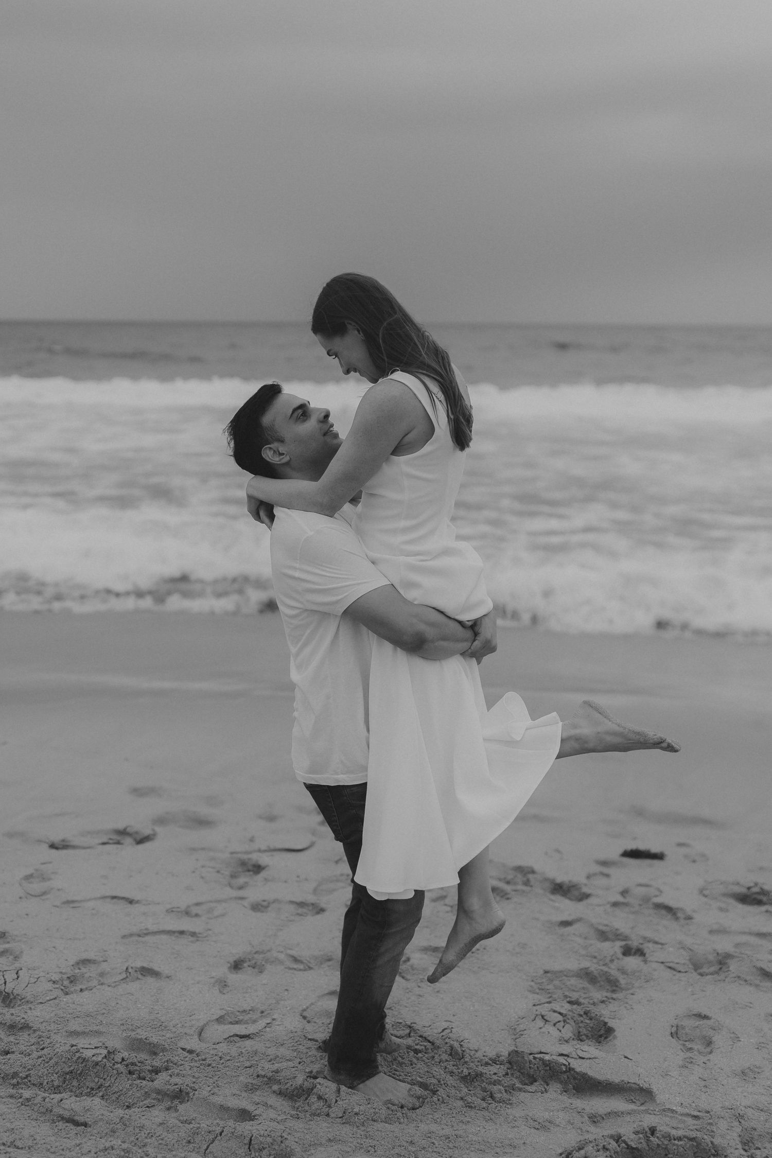 A romantic black and white photo series of a couple dancing and embracing on a beach with ocean waves in the background.