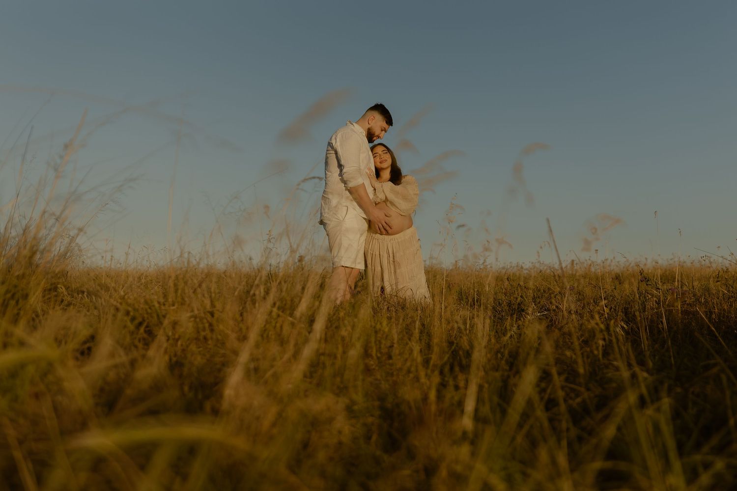 A couple stands together in a golden wheat field during sunset creating a romantic silhouette against the warm sky.
