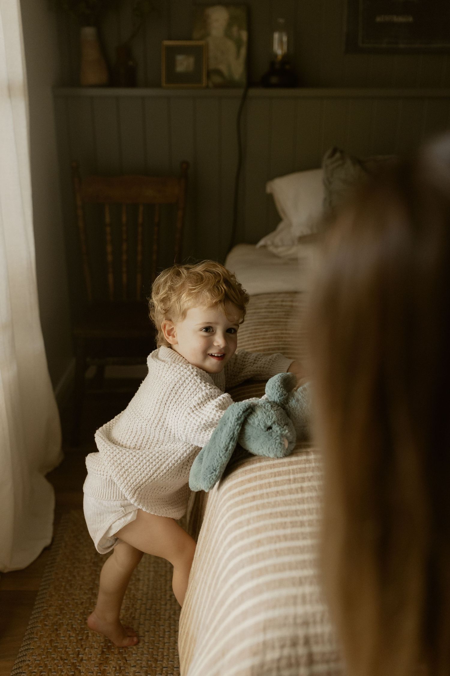 Small child in cream sweater smiling while playing with blue stuffed toy near window.
