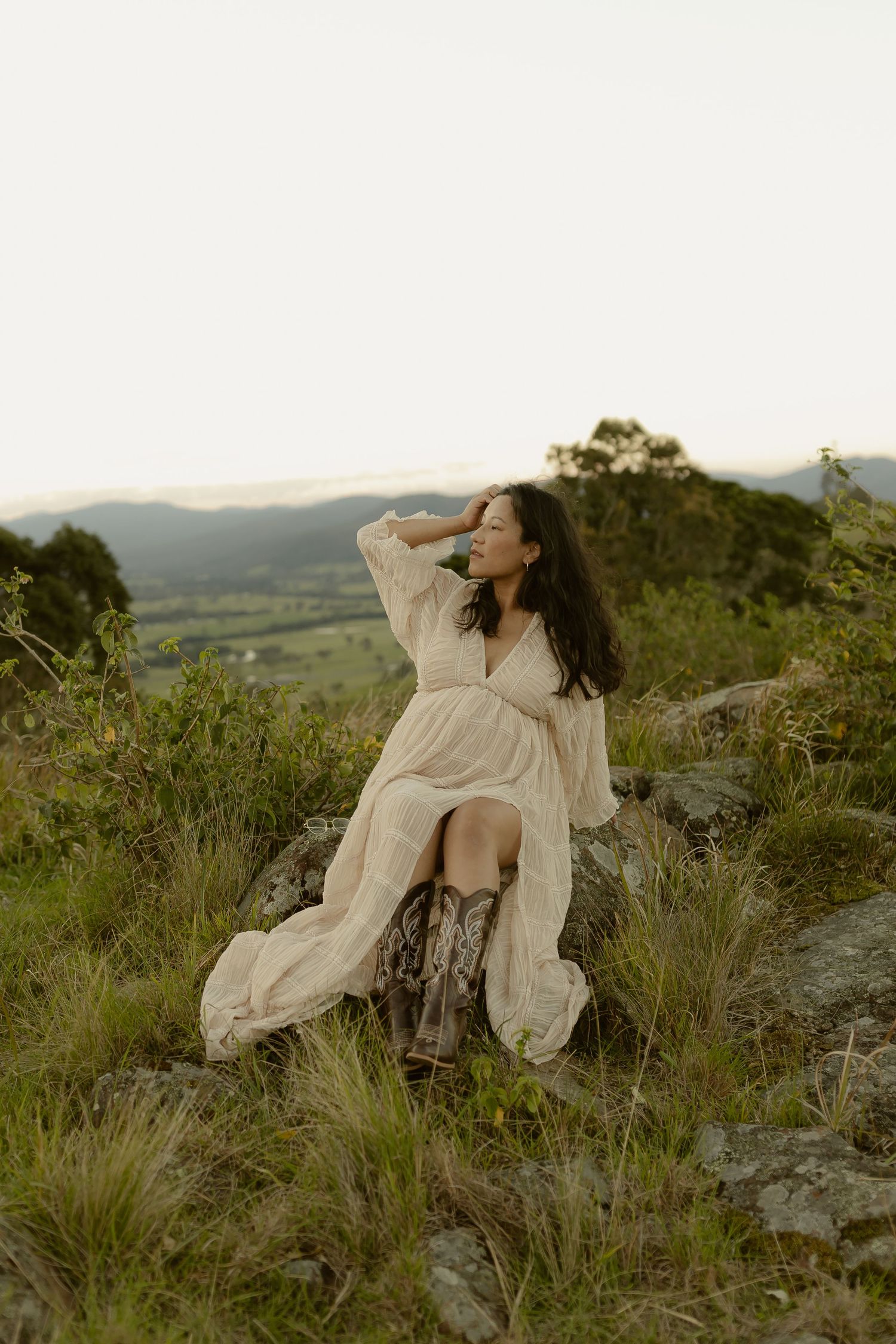 Woman in white dress and cowboy boots sits on rocks in lush green hills at dusk.