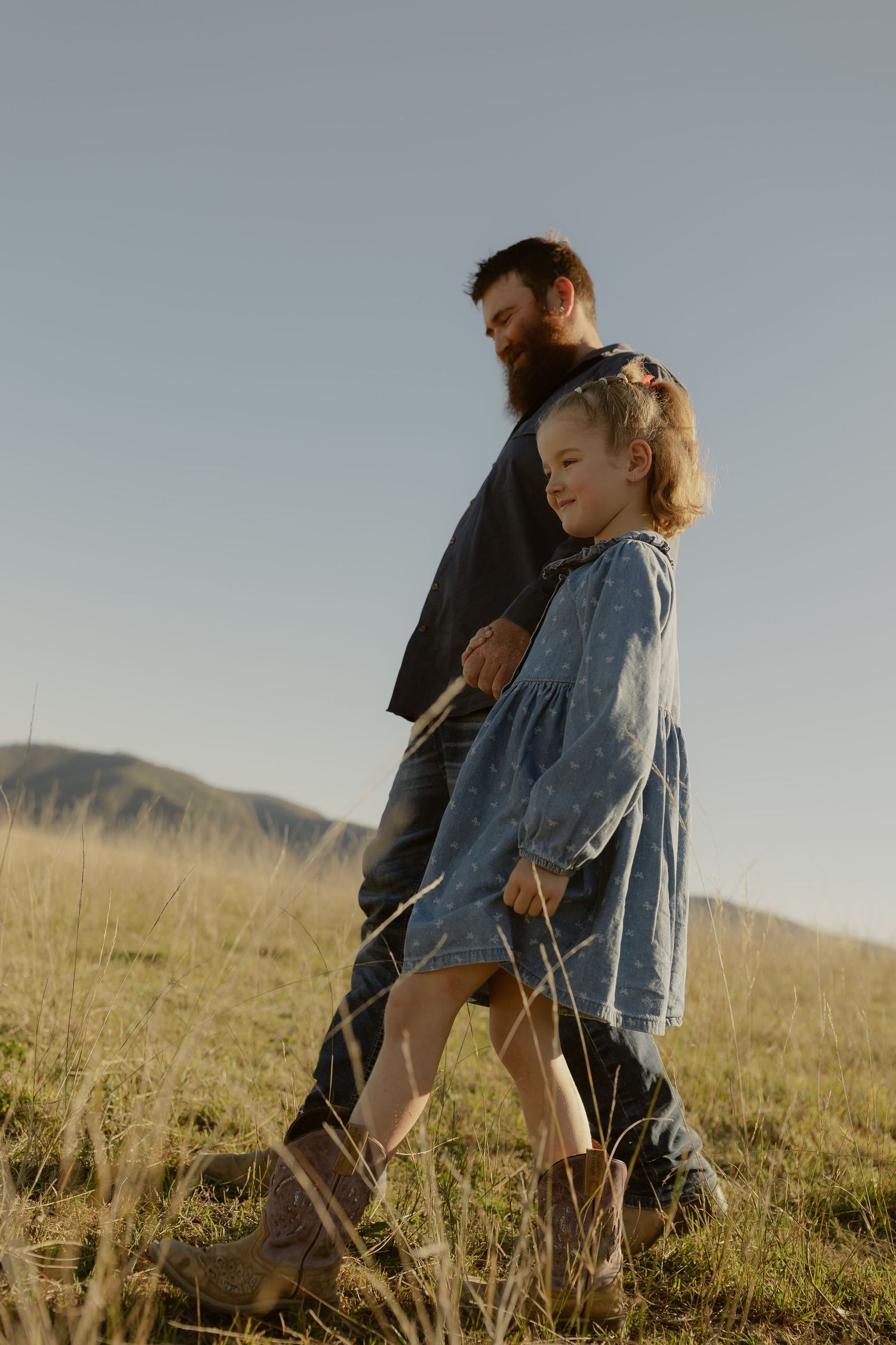 Bearded father walking with young daughter in blue dress through golden grass field with mountain backdrop.