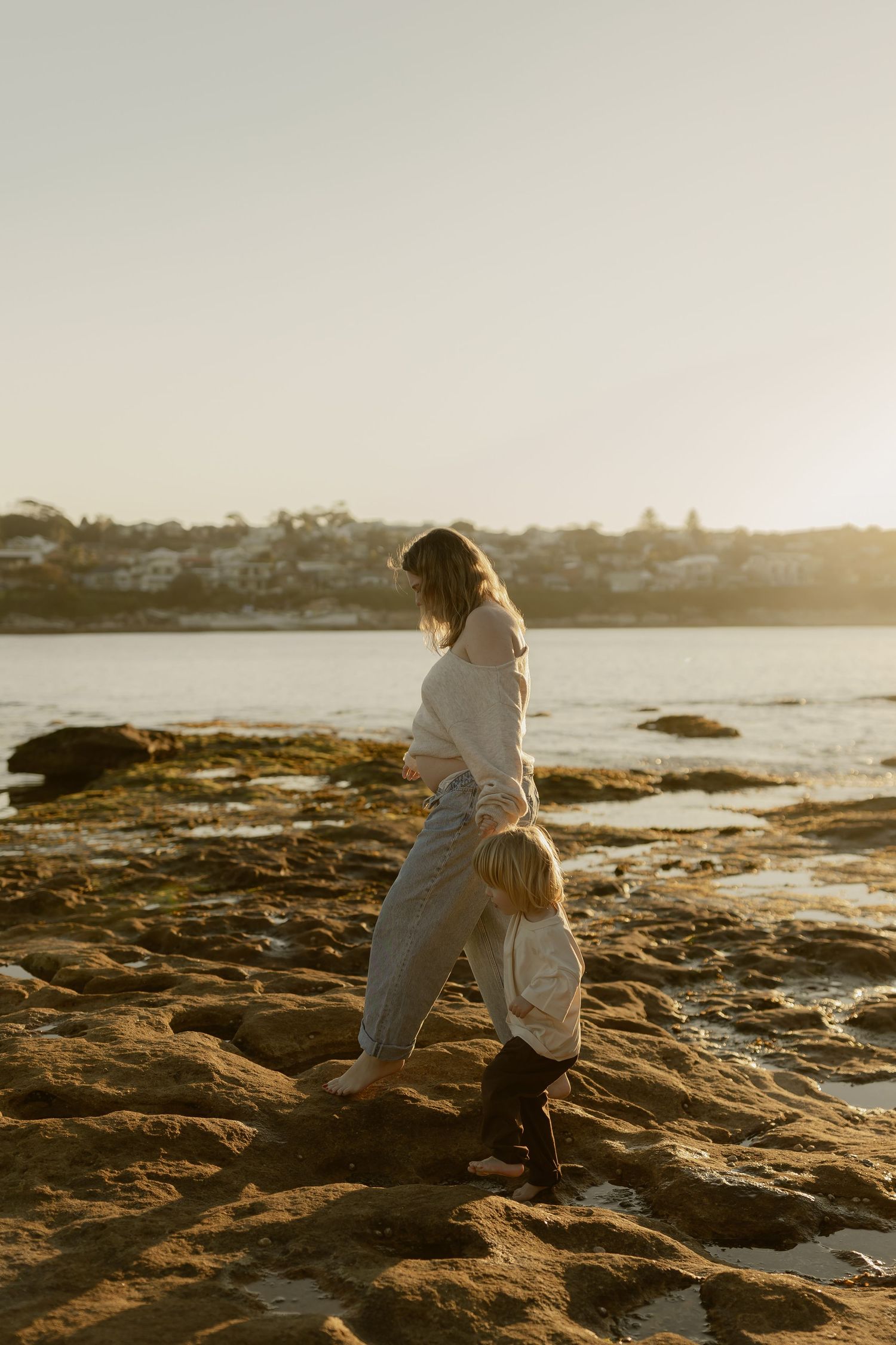 Mother and young child walking on rocky shoreline during warm golden hour sunset.