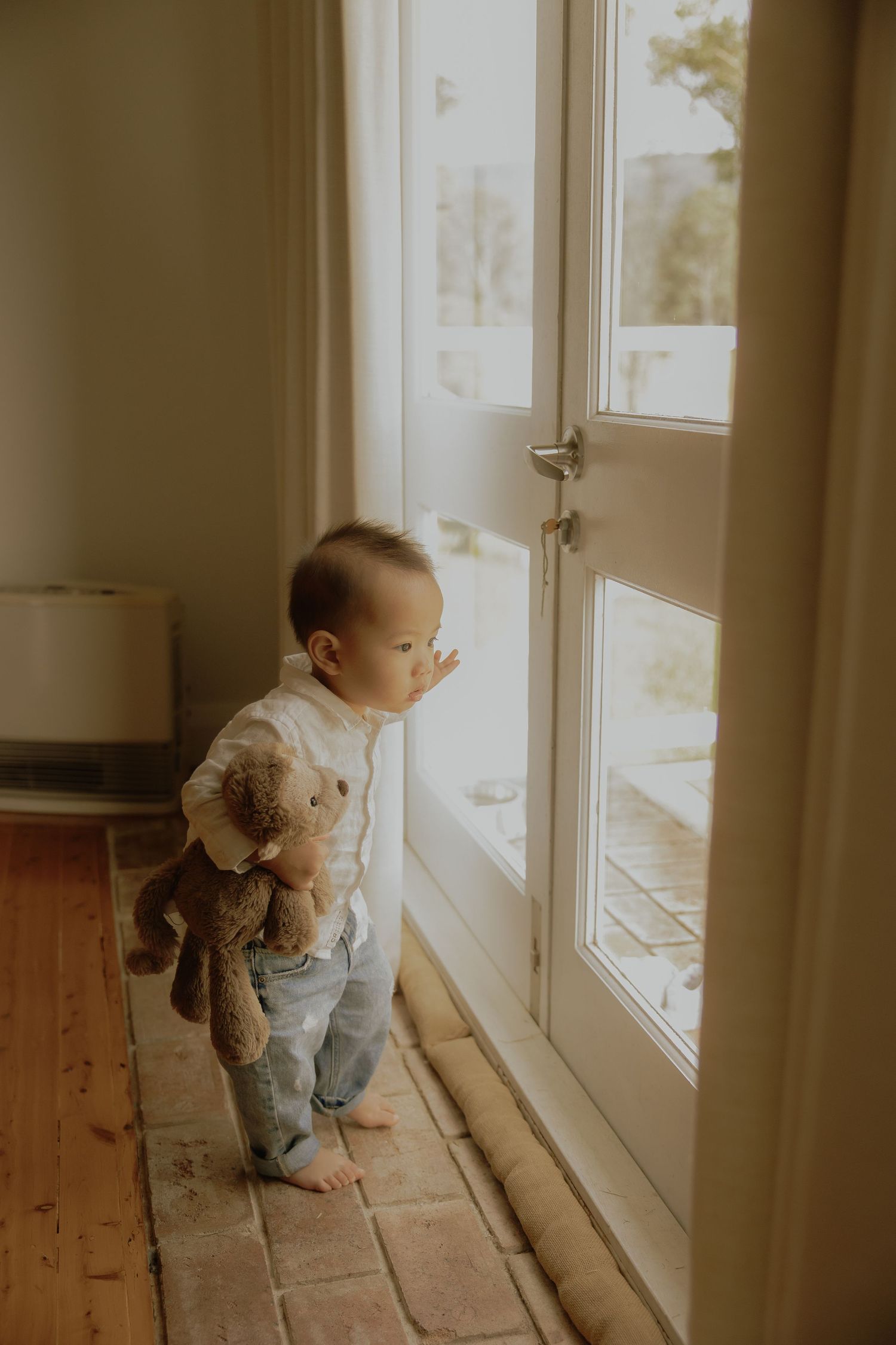 Toddler in white shirt holding teddy bear looking out a glass door indoors.