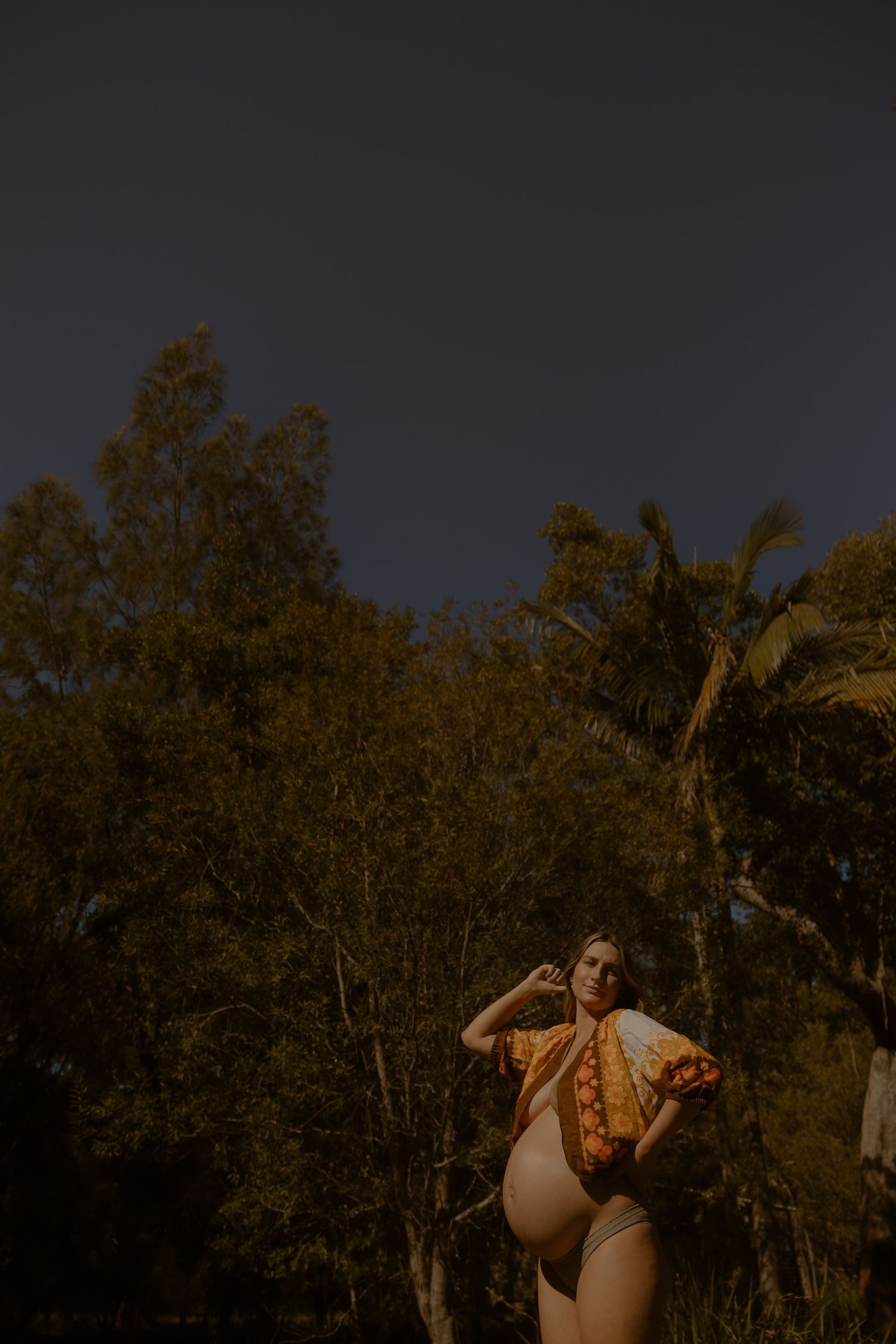 Pregnant woman in bikini and open floral shirt posing outdoors among tropical trees at dusk.