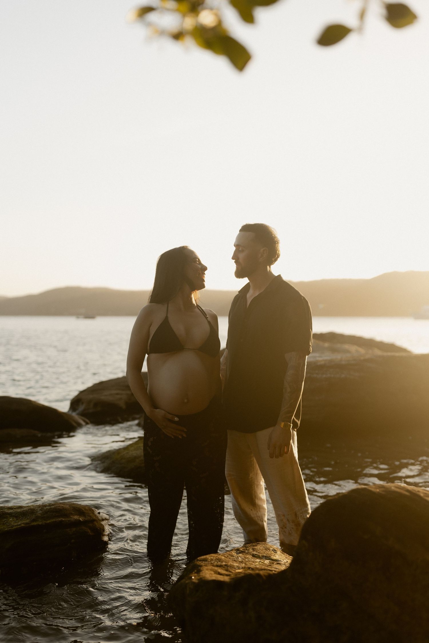 Expecting couple shares intimate moment on rocky shore during sunset, silhouetted against golden water and mountains.