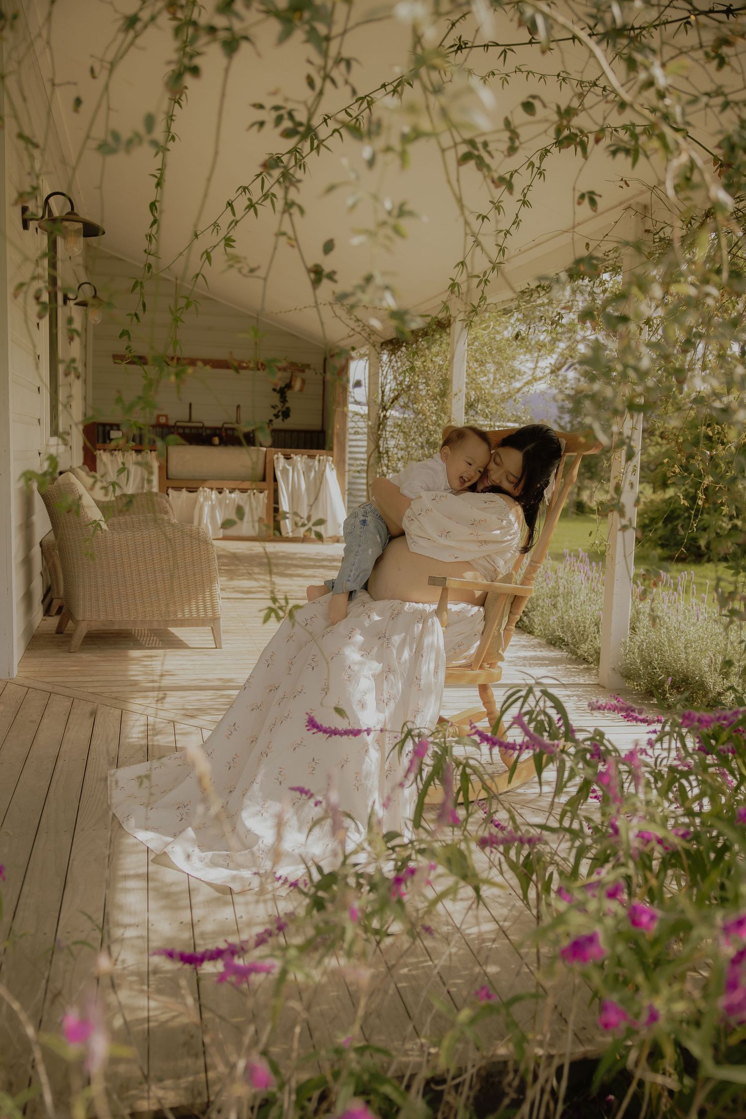 A pregnant woman in a white floral dress sits in a rocking chair on a porch, cradling a child among lush greenery.