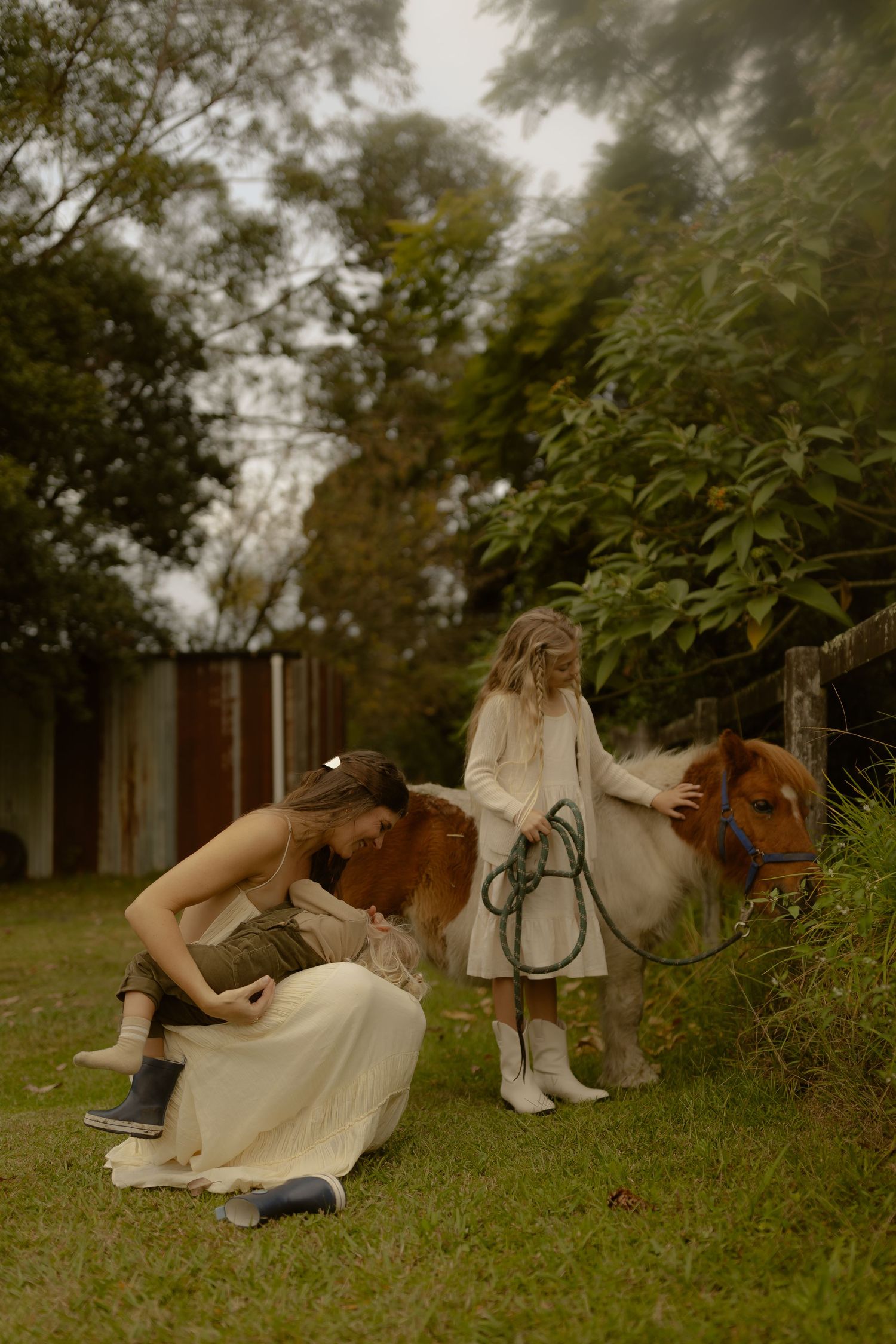 A woman and child interact with a small brown and white pony in a rustic garden setting surrounded by trees.