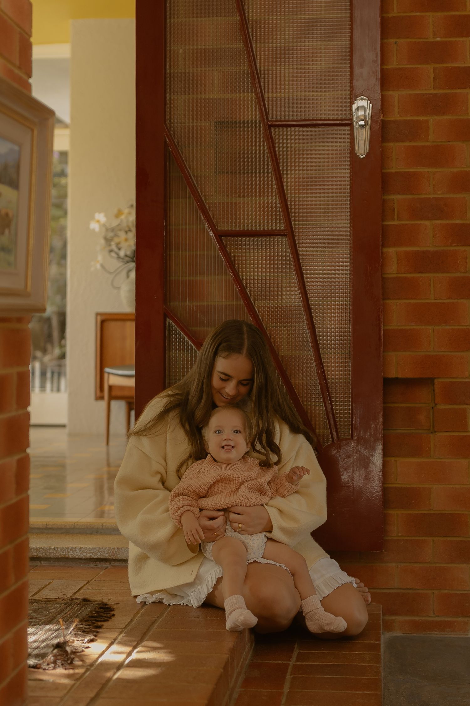 A young woman sits on a tiled doorstep cradling a smiling baby in a pink sweater outside a warm home.