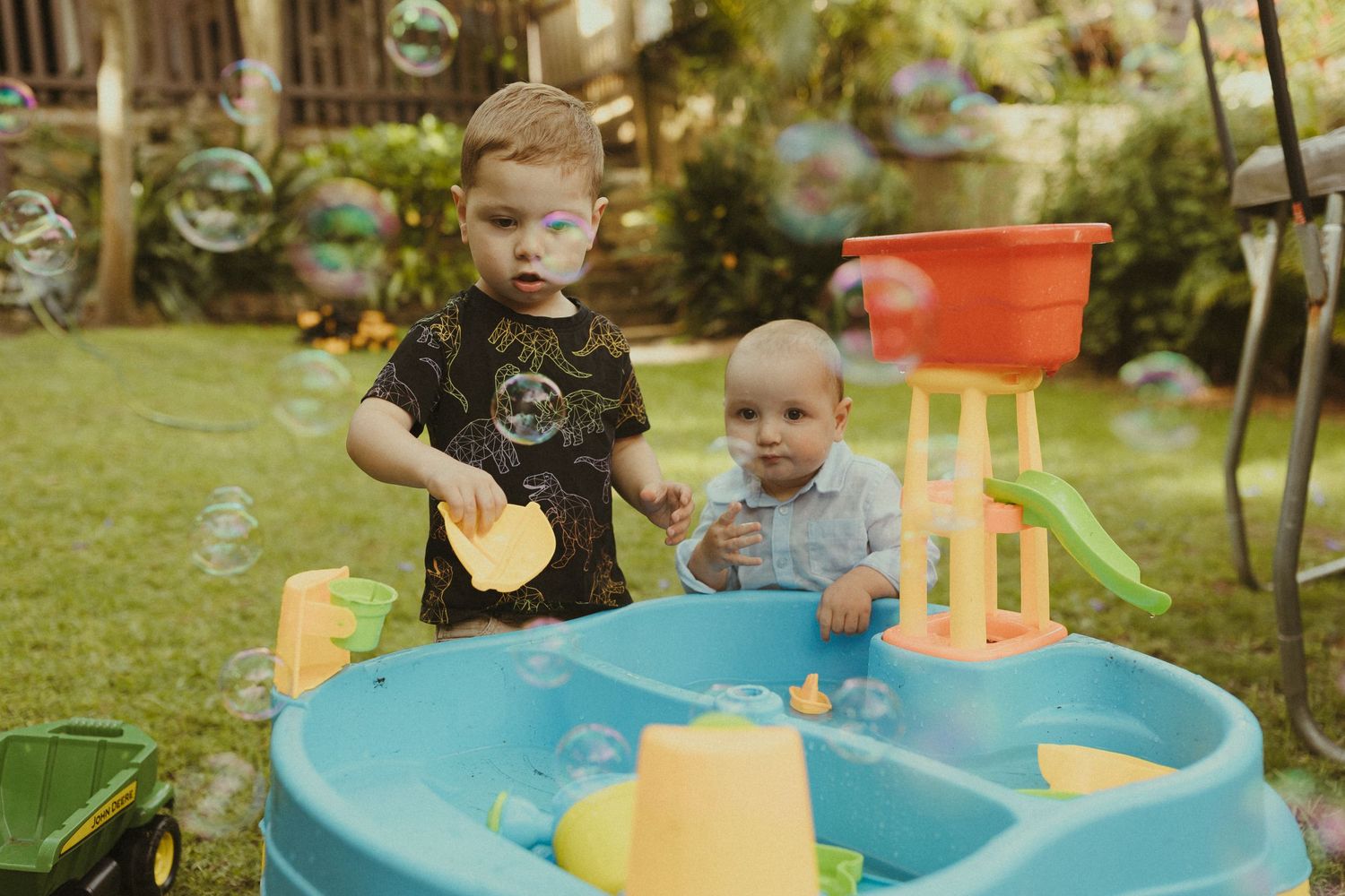 A child plays with bubbles at a blue water table in a sunny backyard setting.