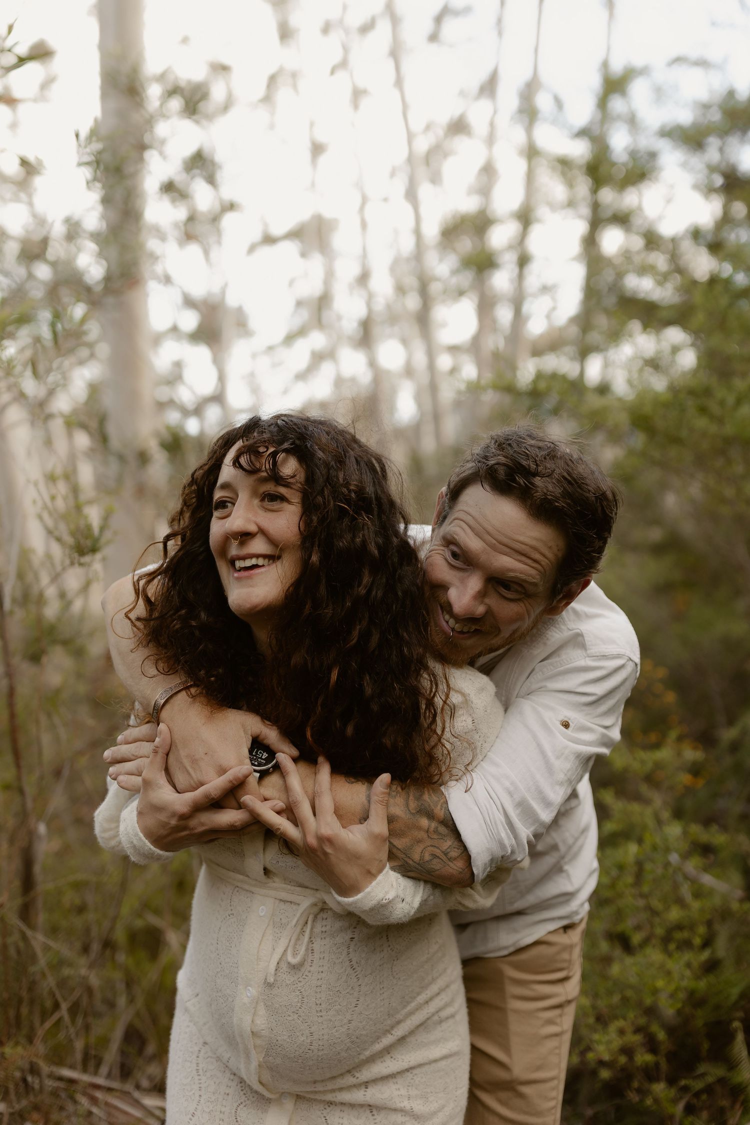A playful couple embraces and laughs together in a wooded outdoor setting during a romantic photoshoot.