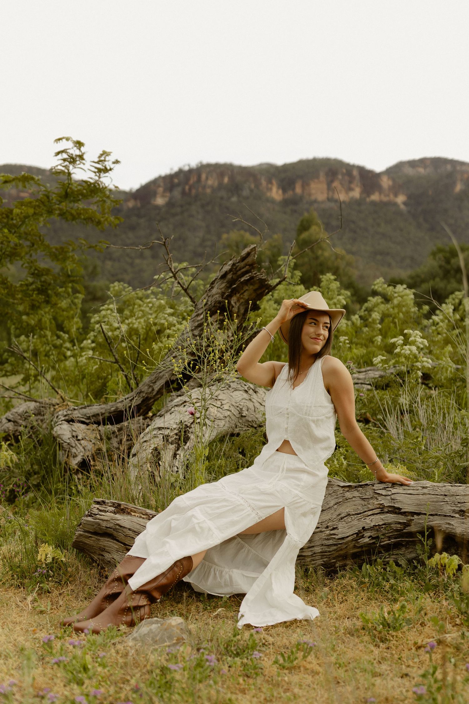 A woman in a white dress reclines on a fallen log in a rustic outdoor setting.