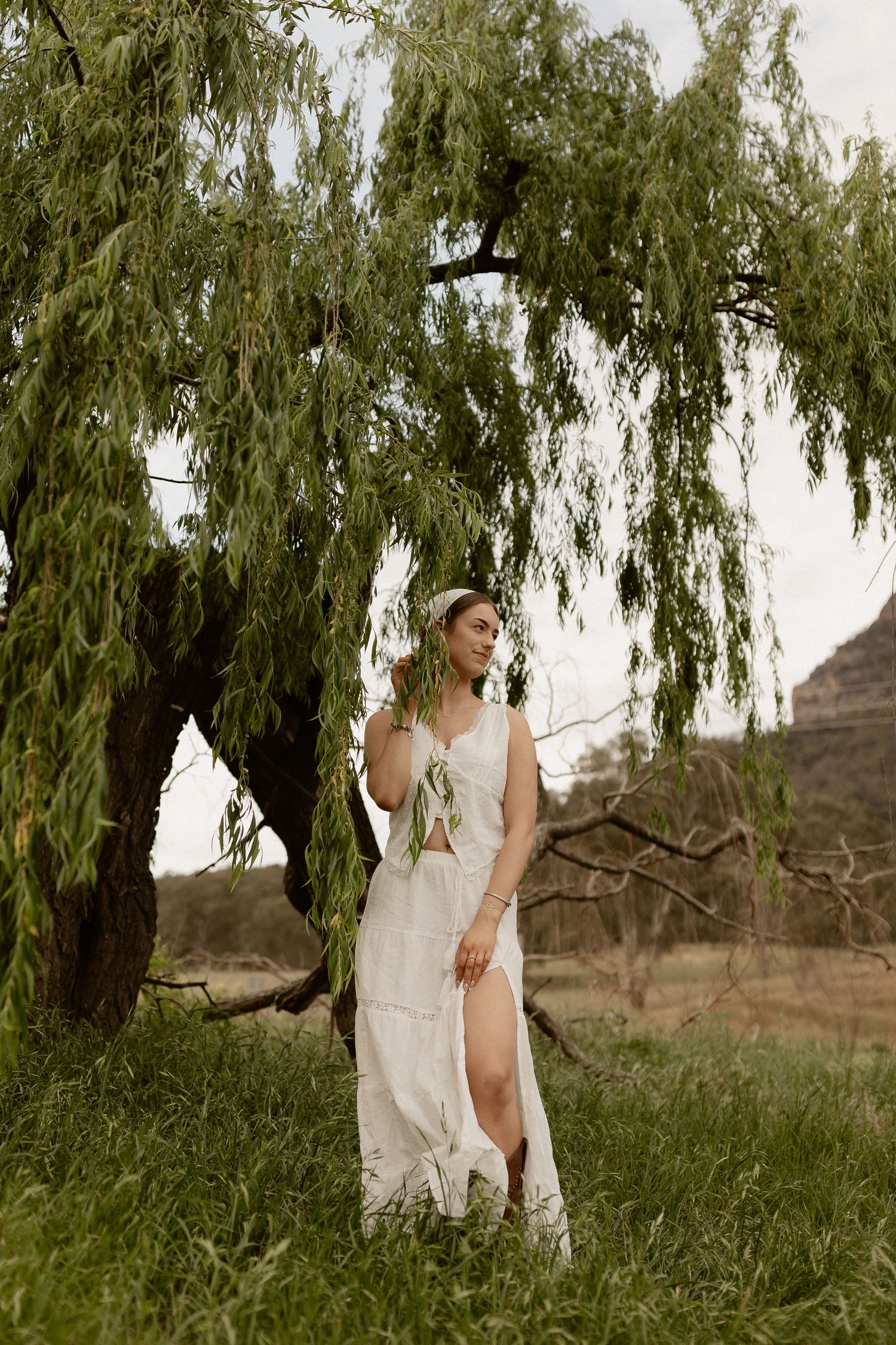 White dress flowing in breeze beneath dramatic weeping willow tree with mountains in background.