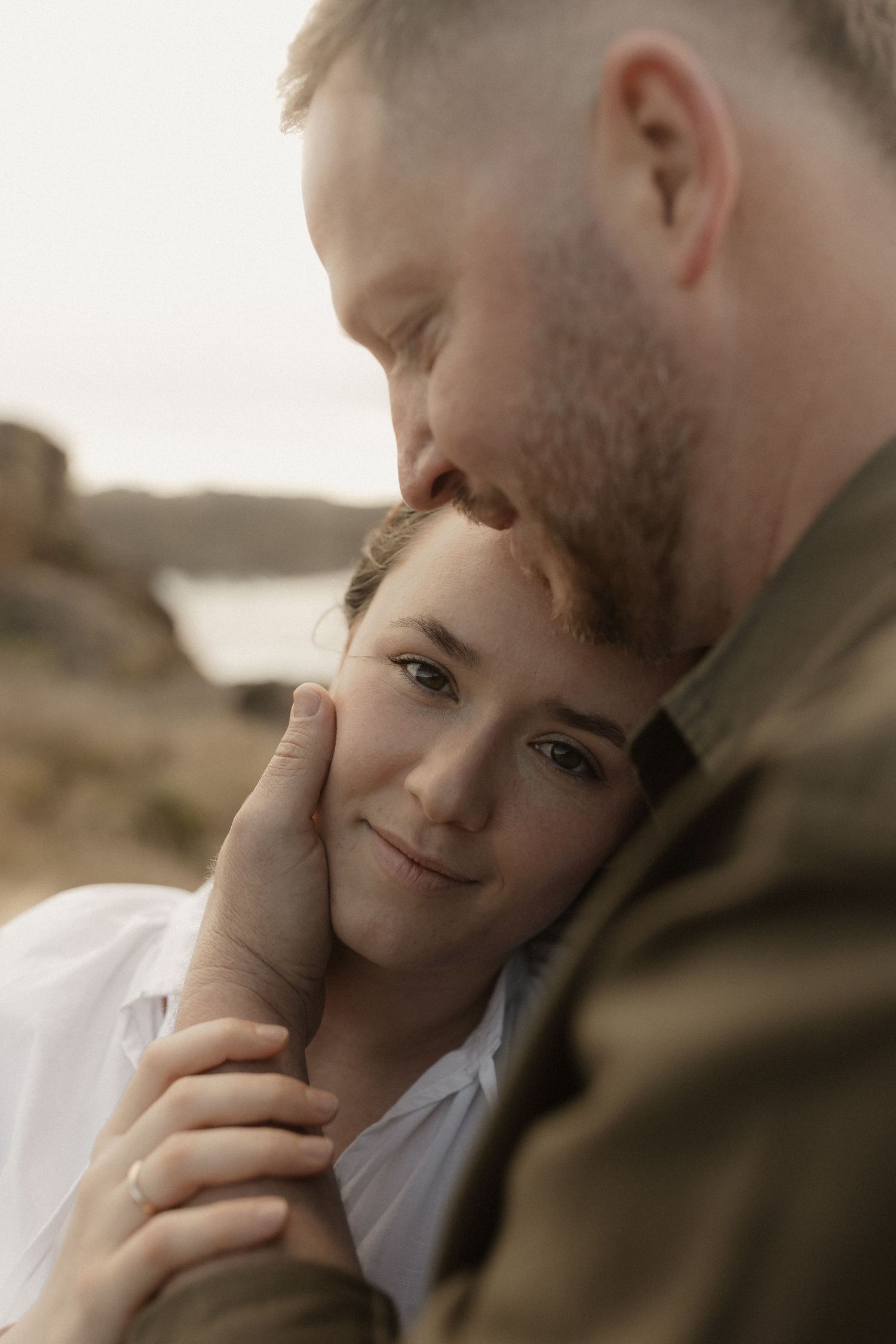 Intimate couple shares tender moment at sunset on rocky coastline with soft warm lighting.