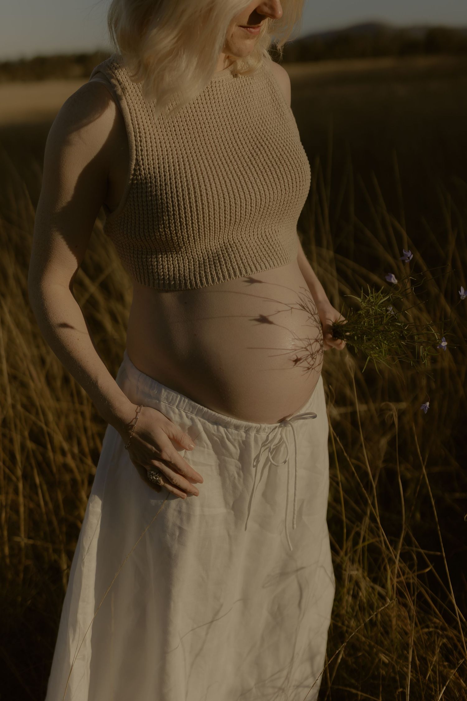 Pregnant woman in beige knit crop top and white skirt standing in golden grass field holding wildflowers.