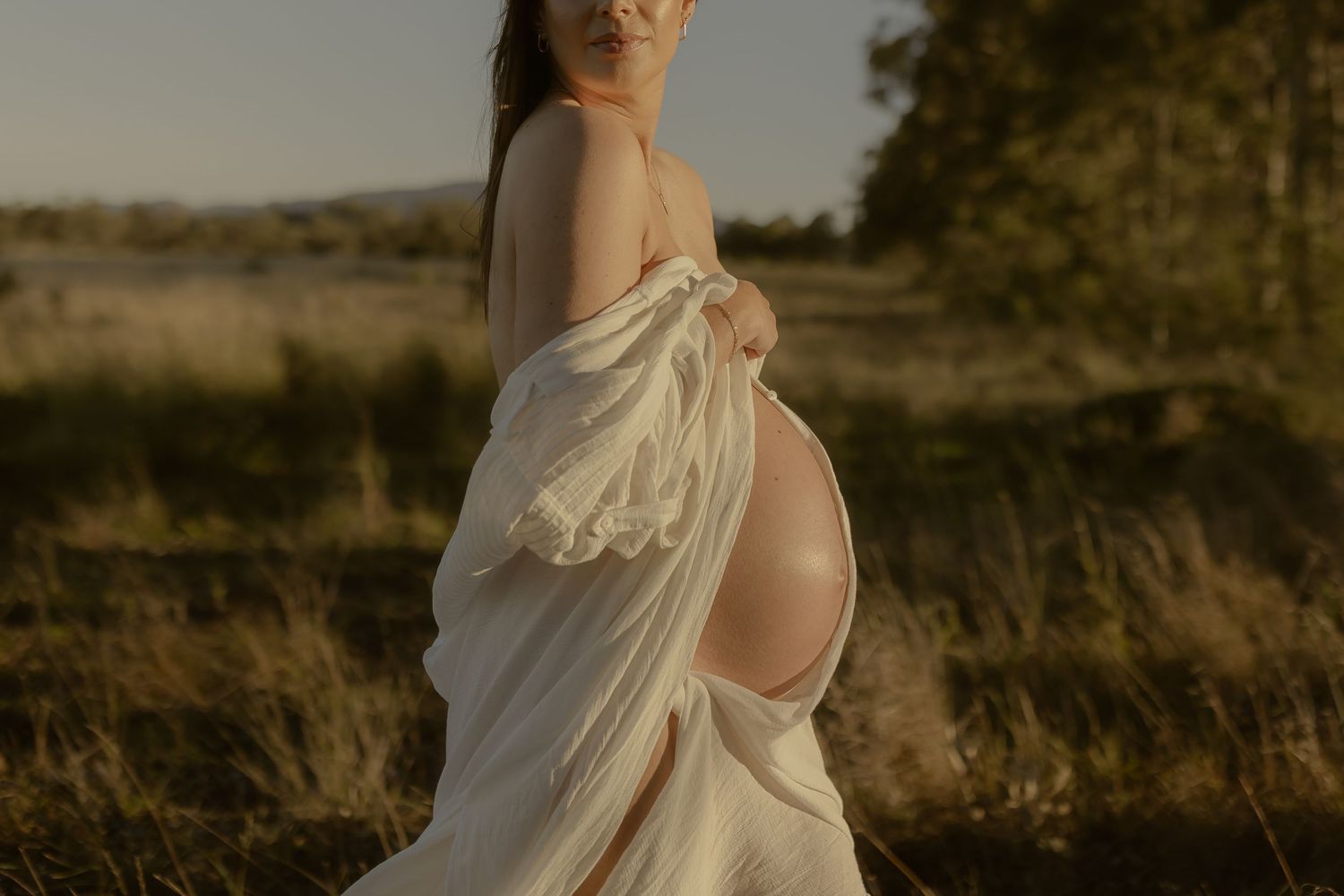 Pregnant woman in flowing white fabric poses outdoors in golden field during sunset maternity photoshoot.