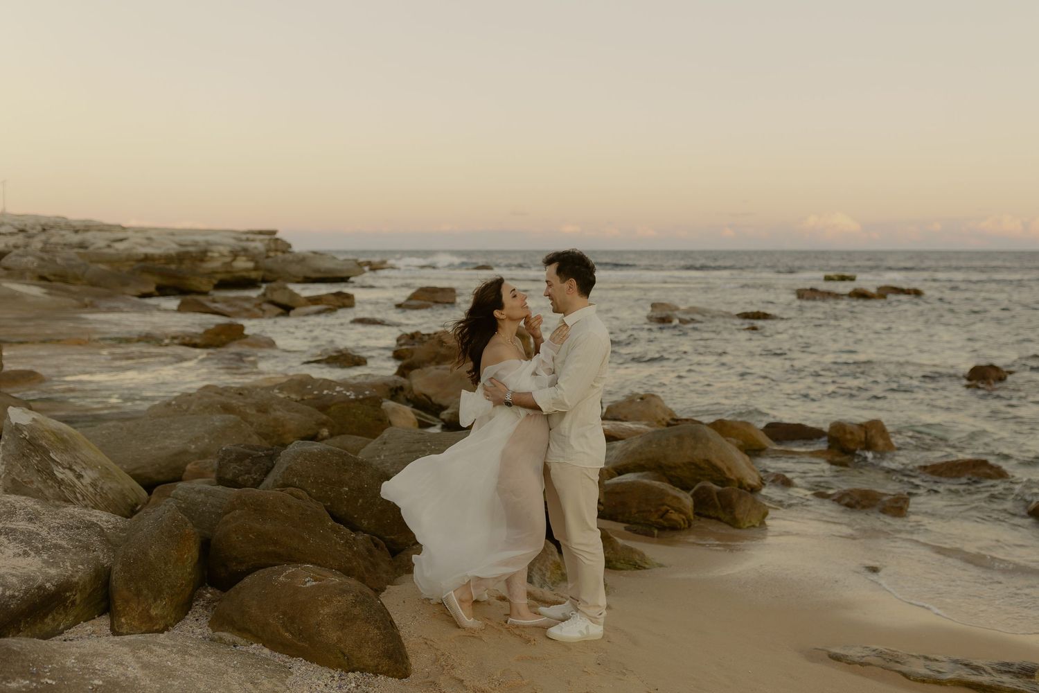 Couple embracing on rocky beach at sunset in wedding attire, waves and golden sky in background.