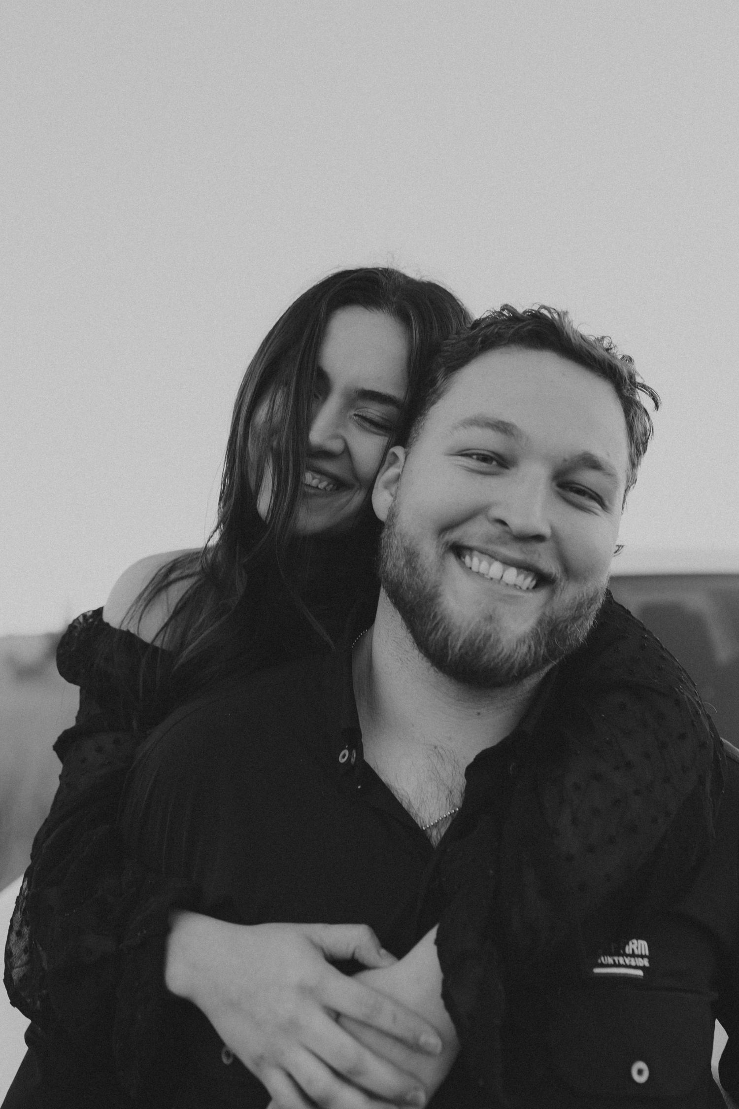 Black and white photo of woman hugging man from behind, both smiling warmly in an outdoor setting.