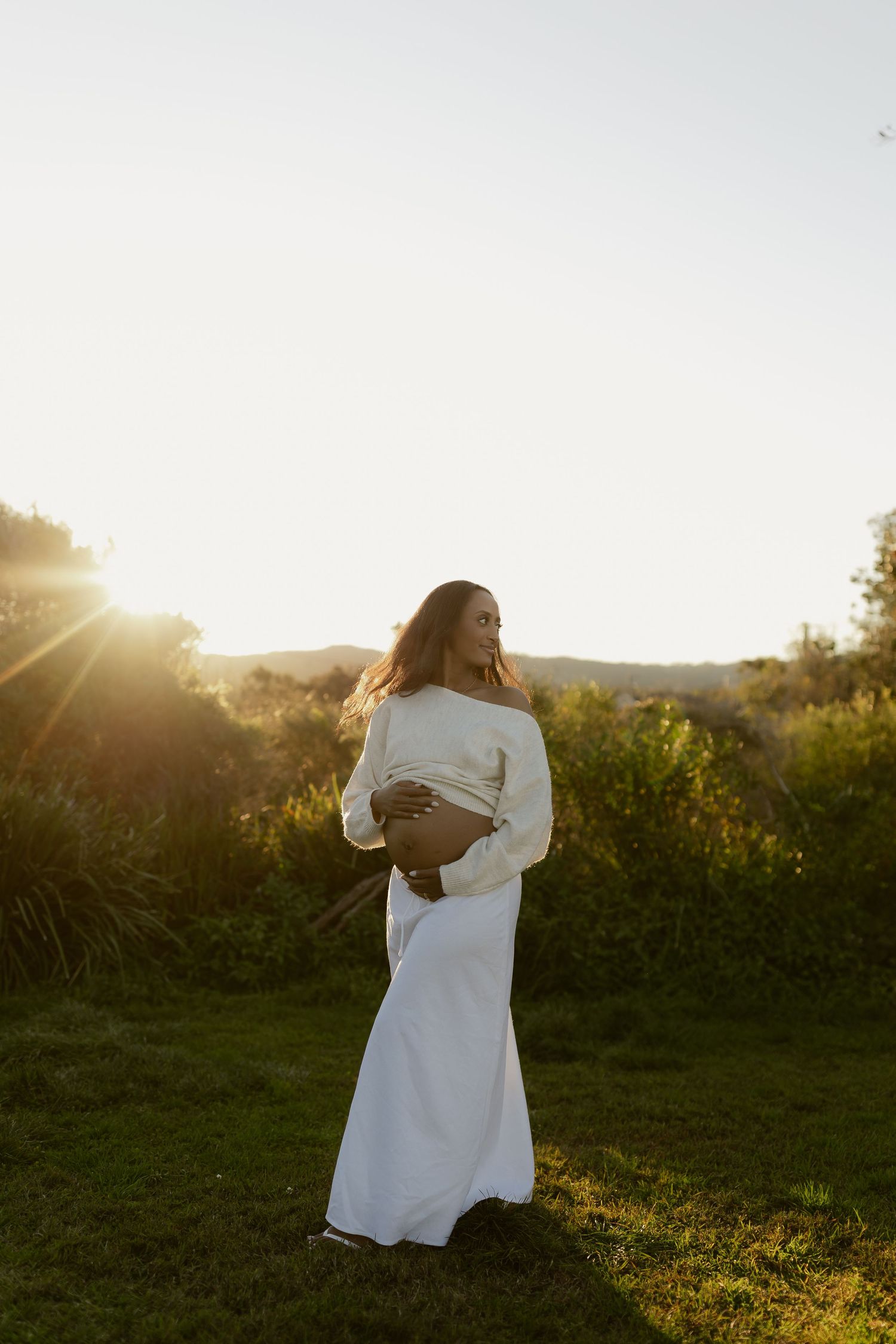 Pregnant woman in white dress and sweater cradling baby bump during golden hour in nature.