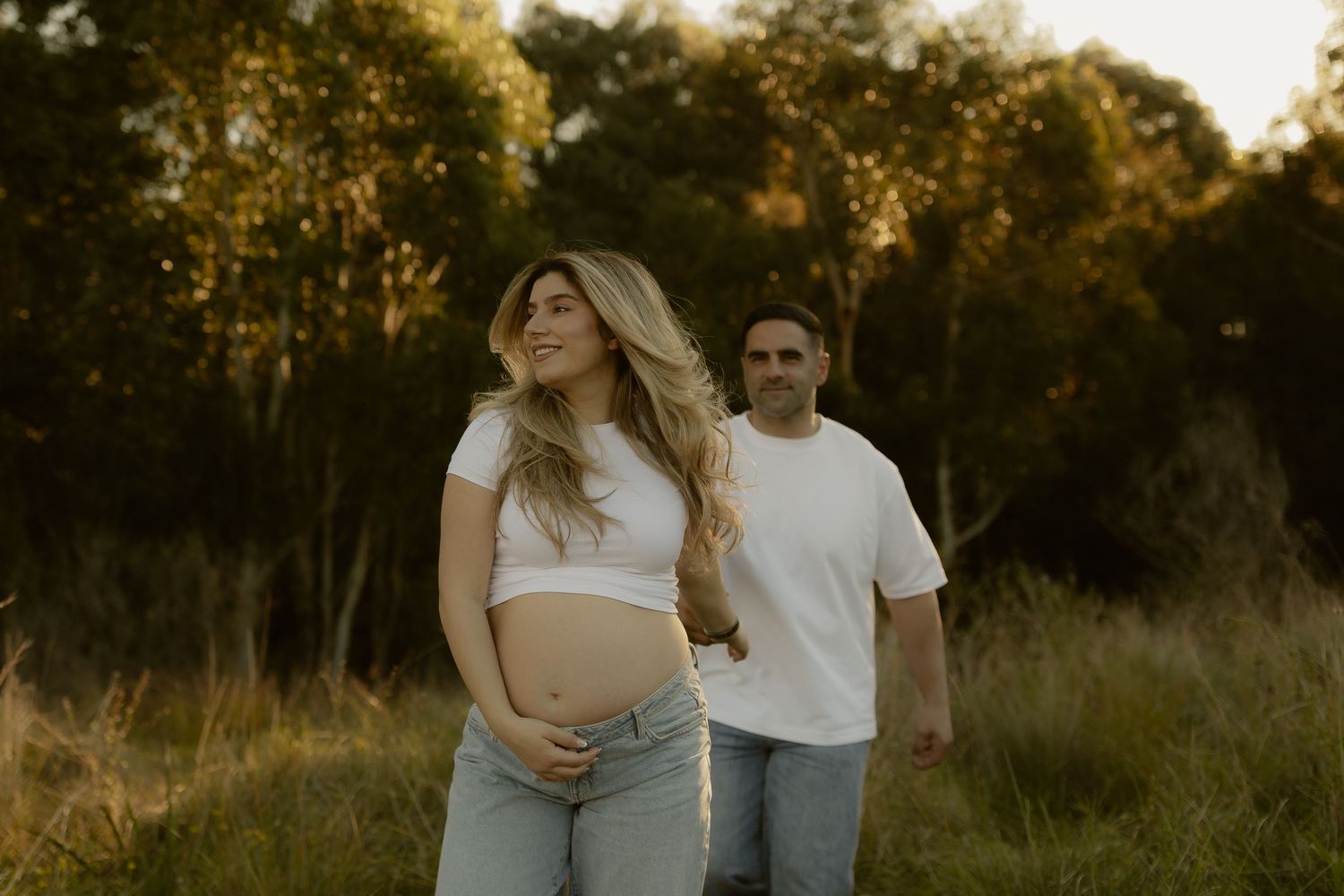 Pregnant woman in white crop top and jeans holds baby bump outdoors at golden hour with partner standing behind her.