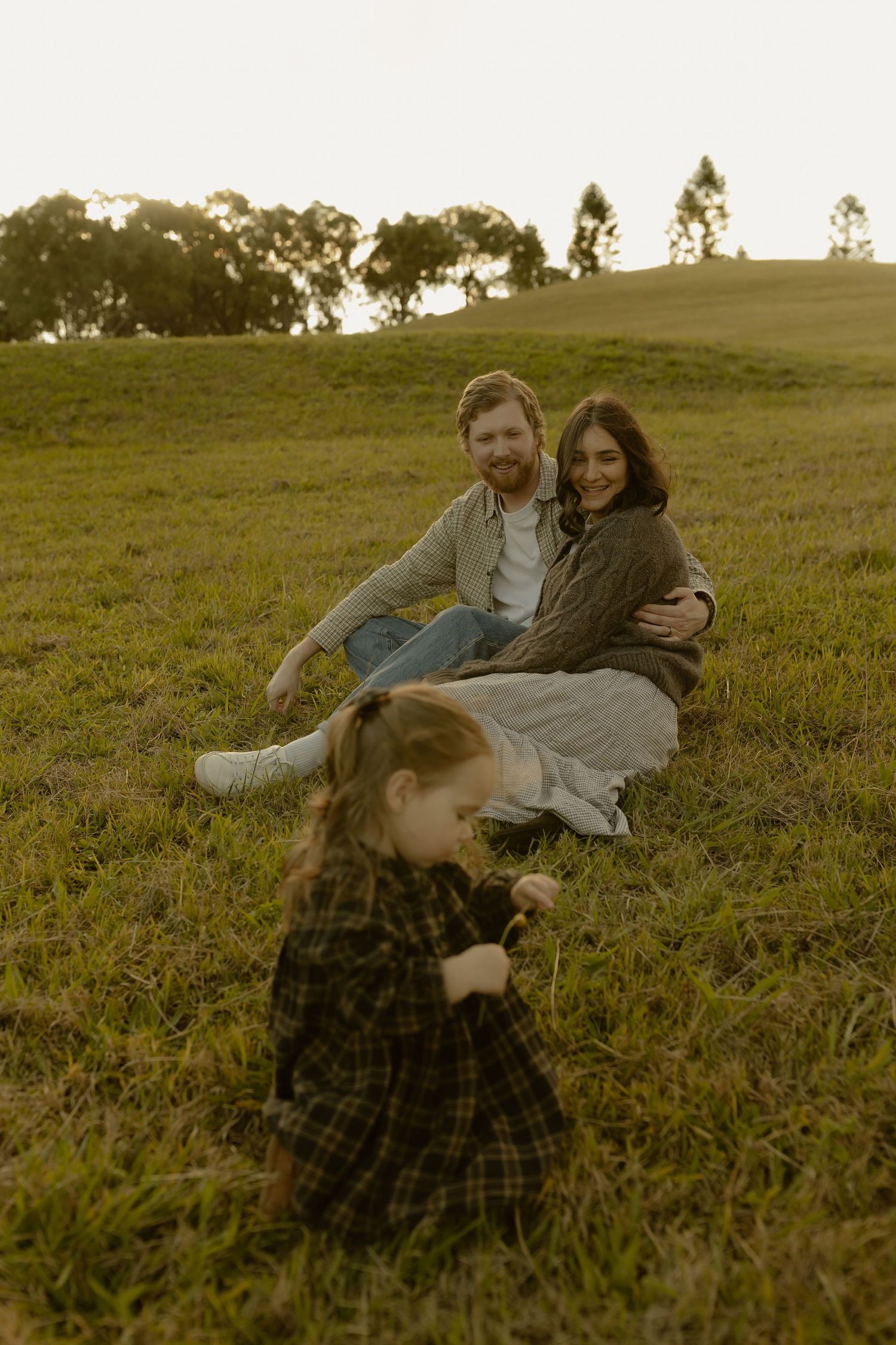 A couple sits on a green grassy hill smiling as their young daughter in a plaid dress plays nearby.