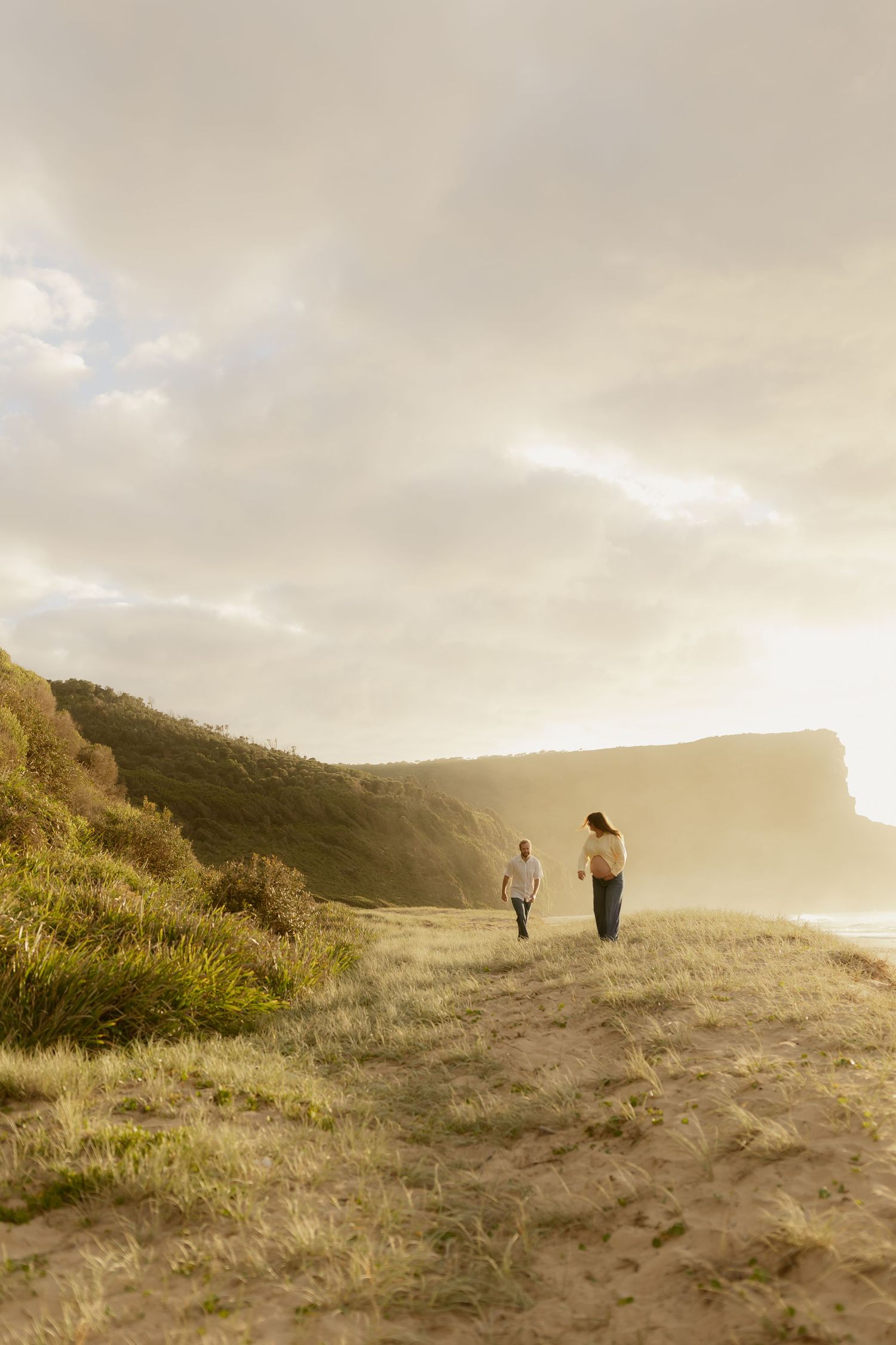 Two people walk along a misty hilltop path overlooking the ocean during golden hour.