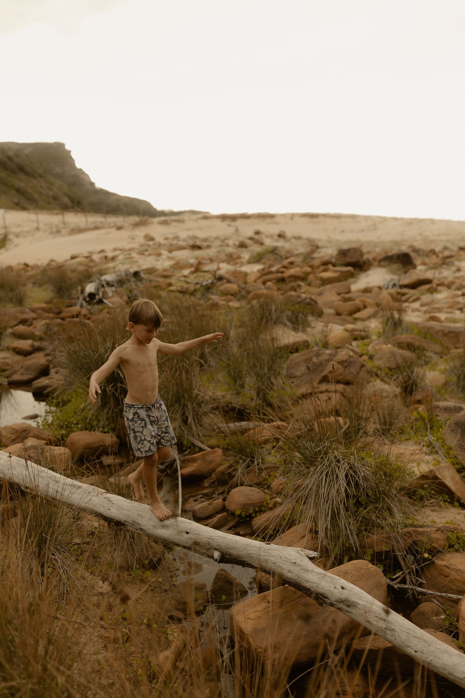 Child balancing on a log while exploring rocky coastal terrain at sunset.