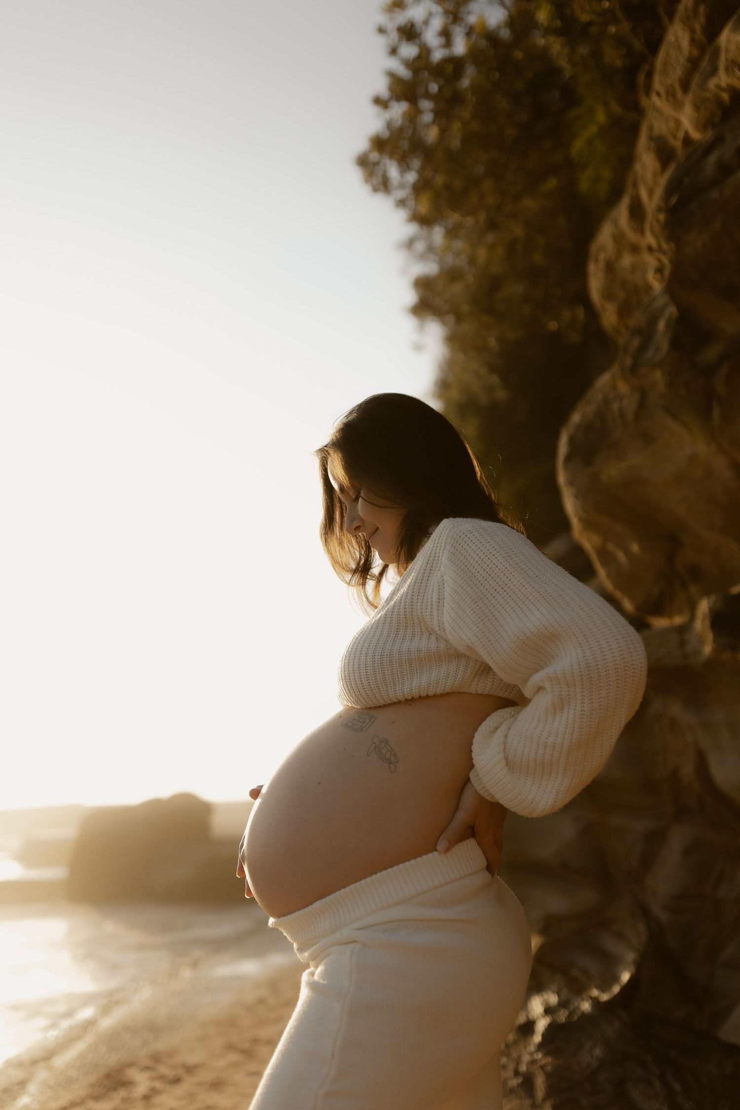 A person in a white sweater cradles their pregnant belly while standing on a beach at sunset.