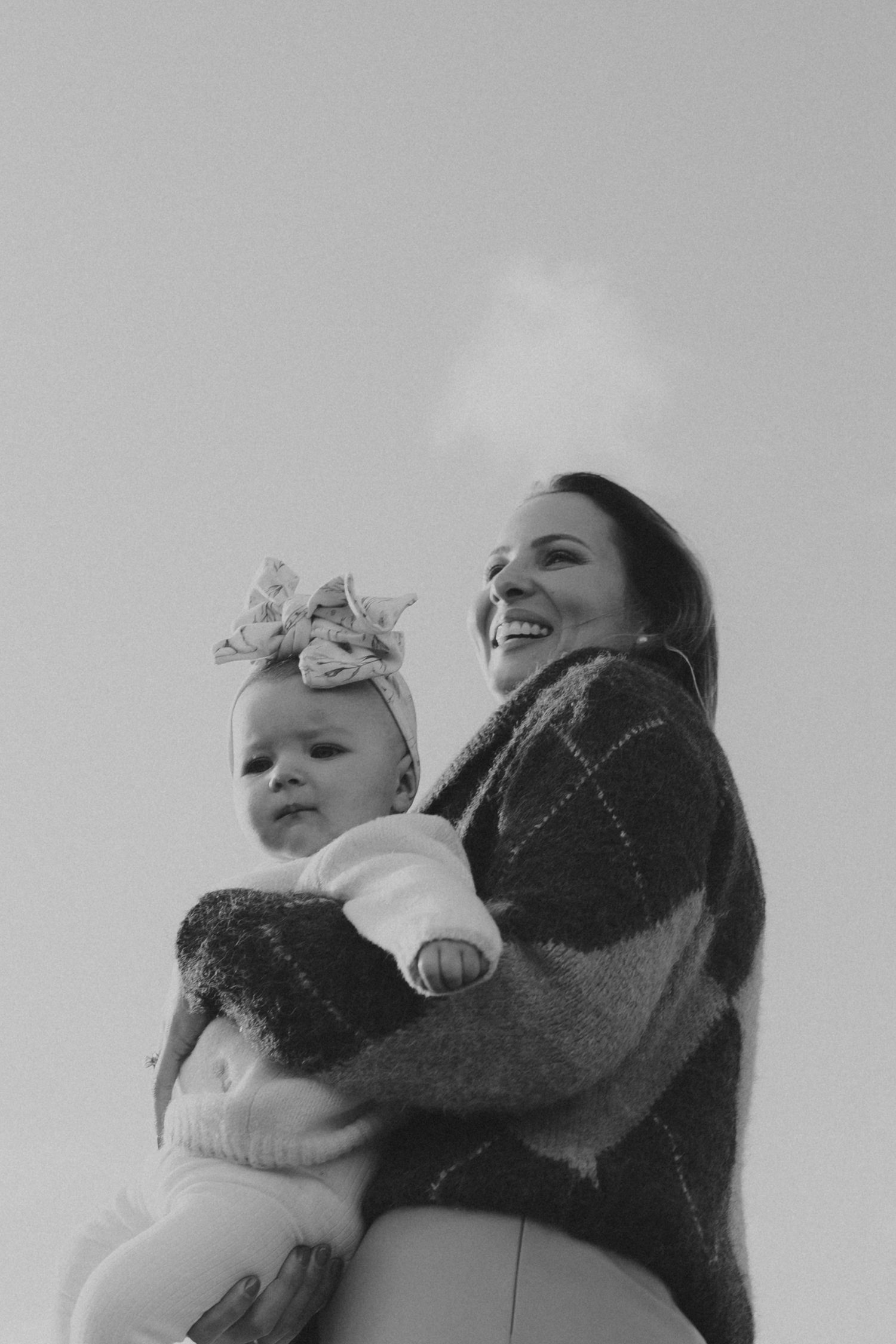 A smiling mother holds her baby girl with a floral headband in a black and white outdoor photo.