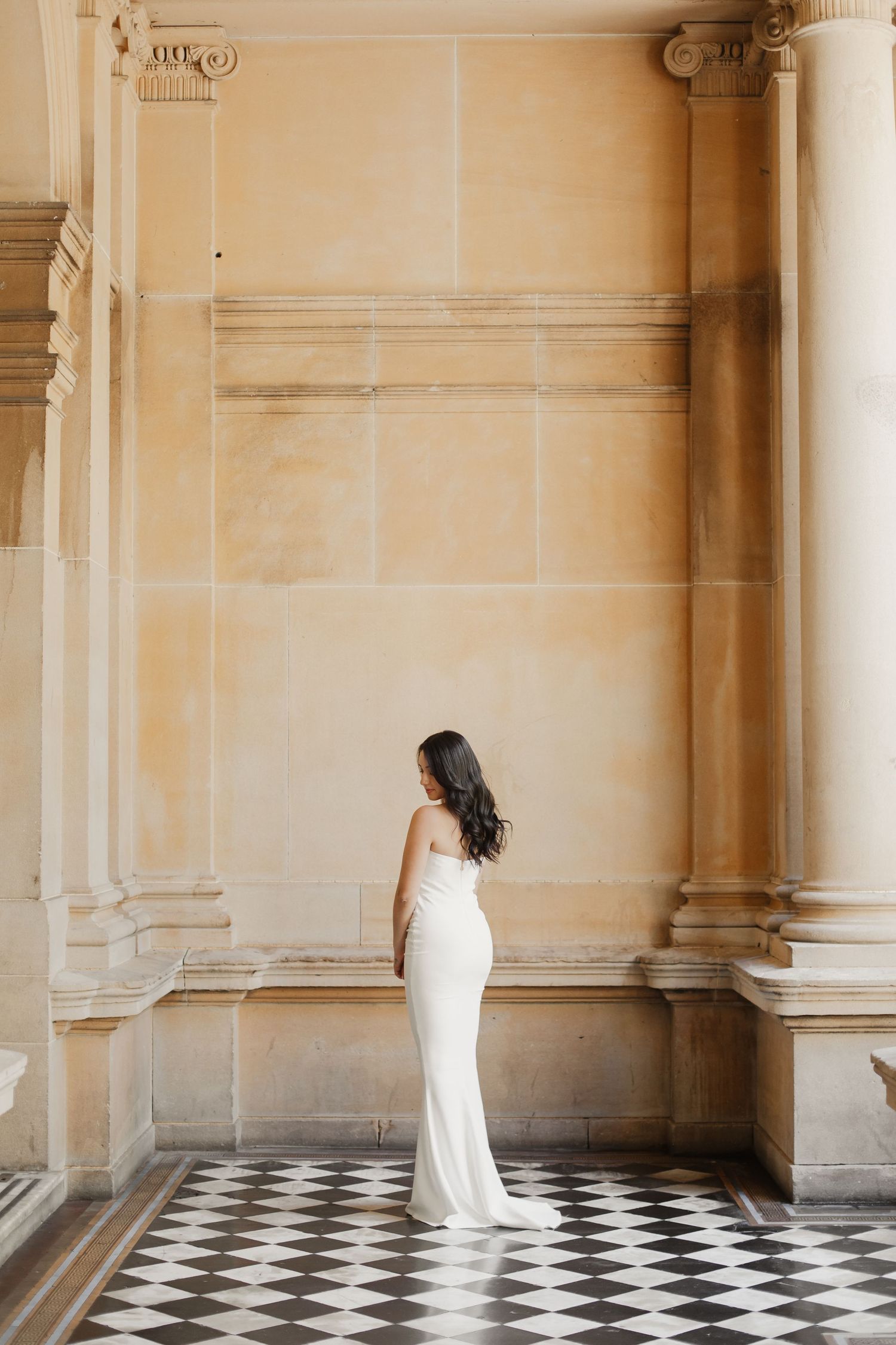 Bride poses in fitted white gown against classical architecture and checkered floor.