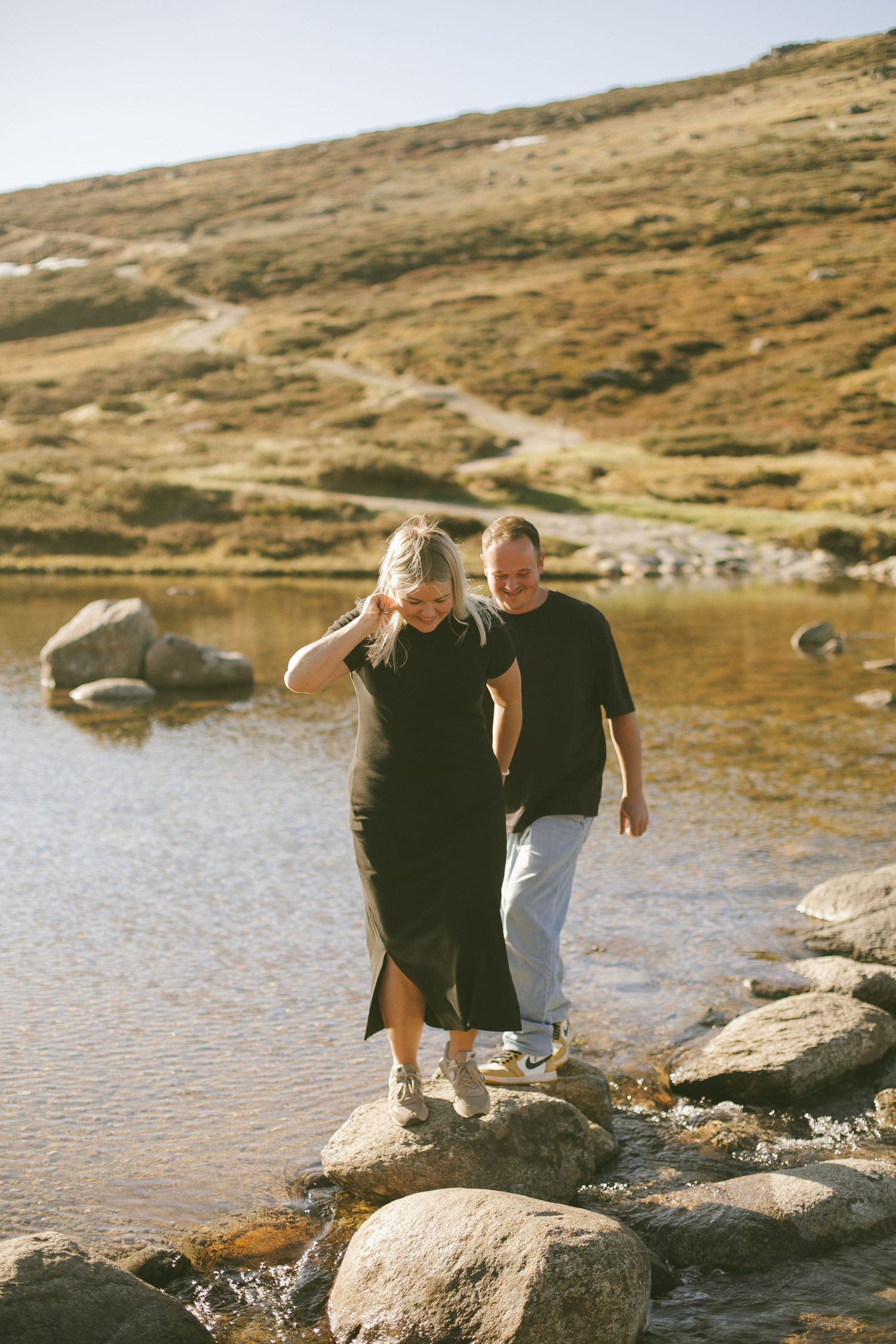 Two people in dark clothing walking together on rocks along a serene mountain lake on a sunny day.