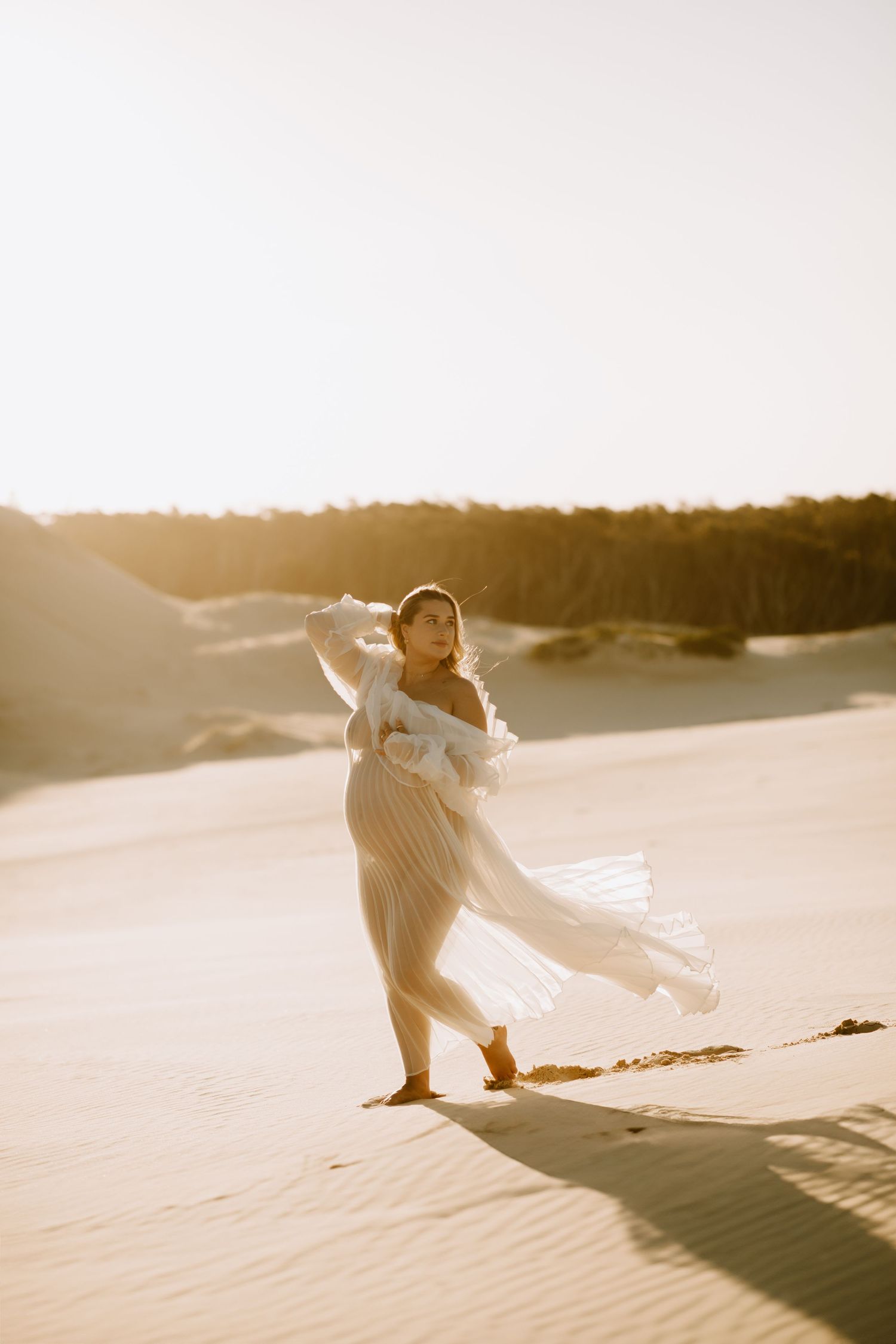 Dreamy desert maternity portrait with flowing white fabric catching sunlight at sunset.