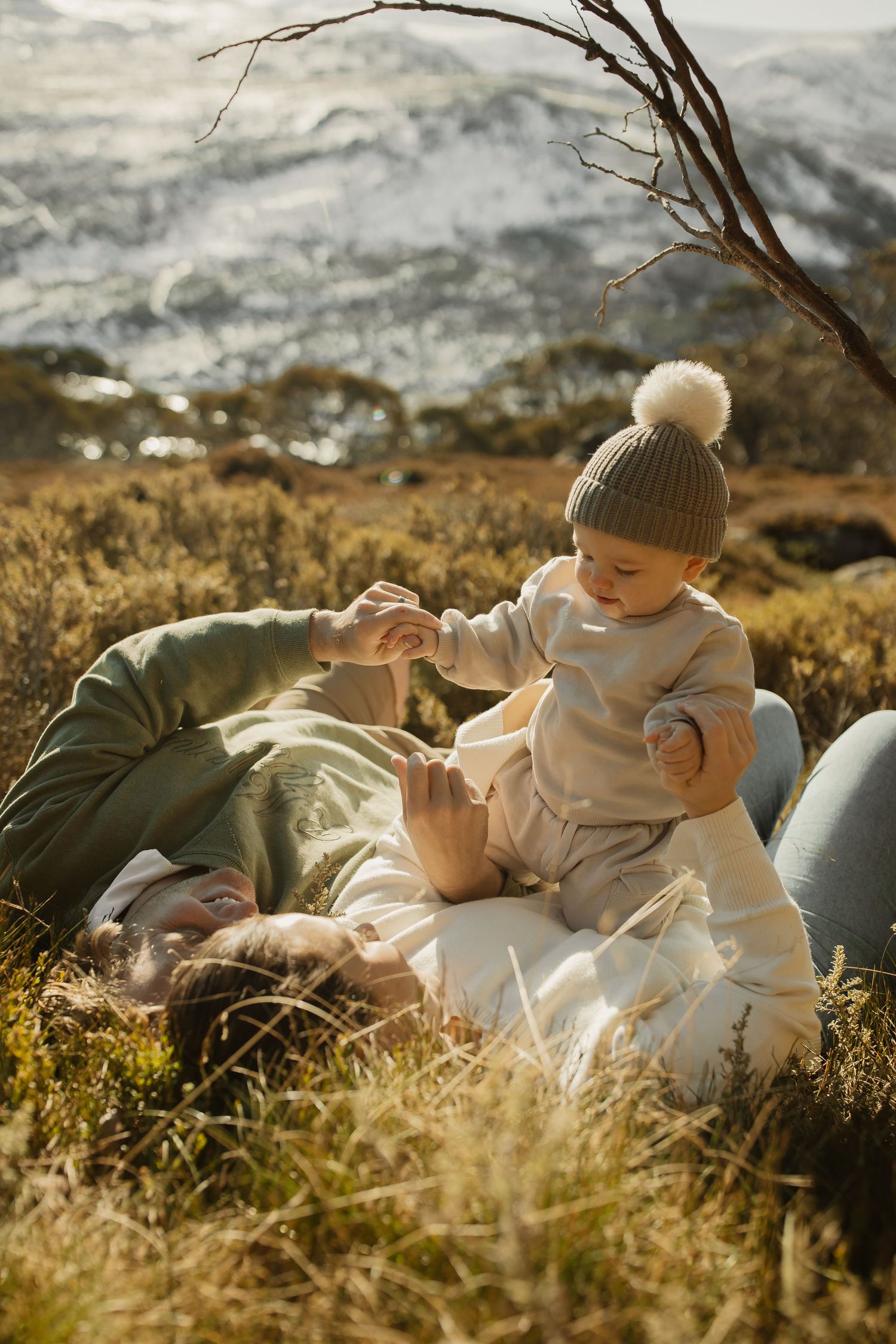 A baby in a knit hat plays while laying in tall grass with adults during a golden hour photoshoot.