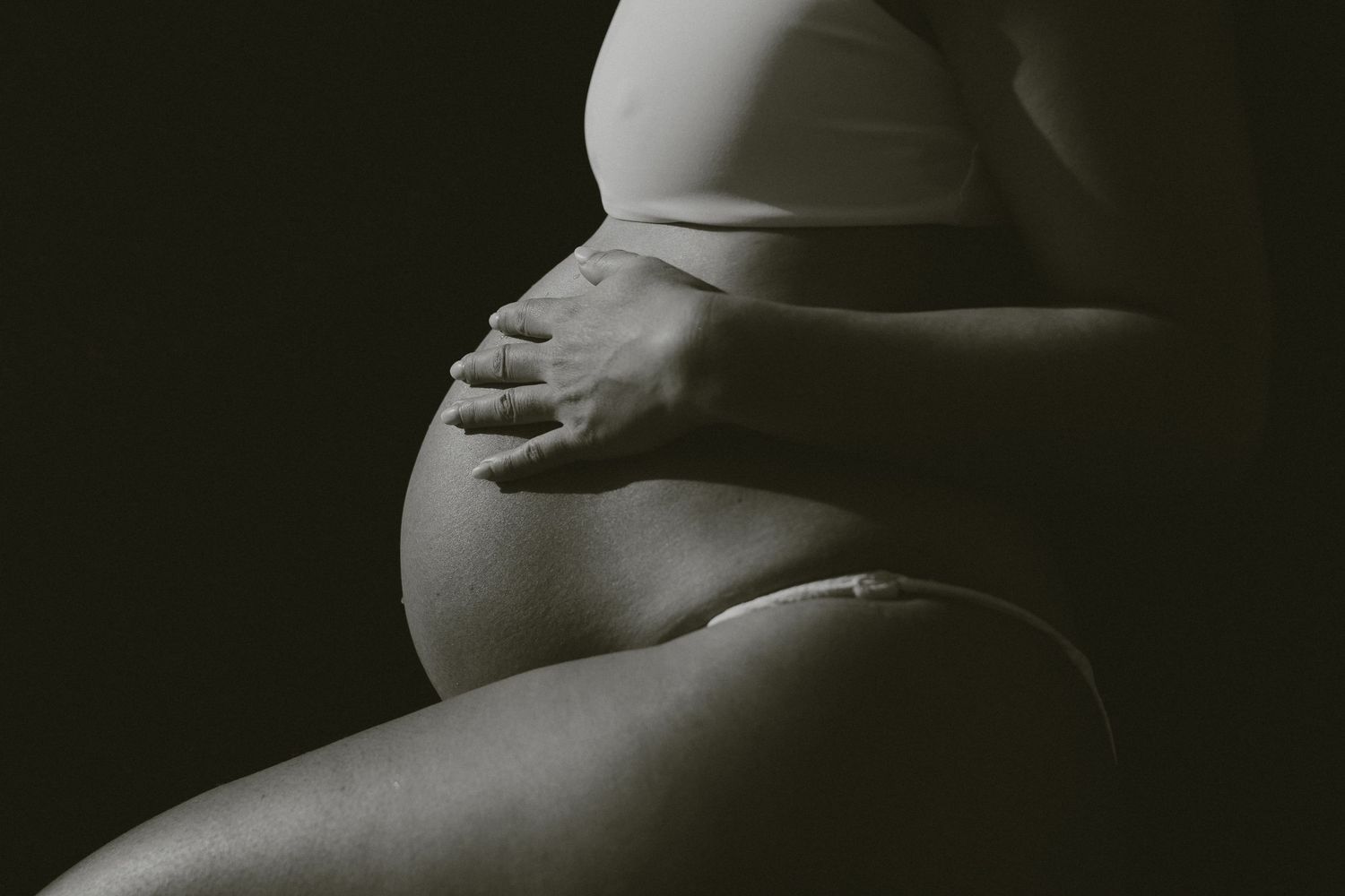 A black and white artistic photograph of a pregnant woman's belly being cradled by hands in a dark setting.