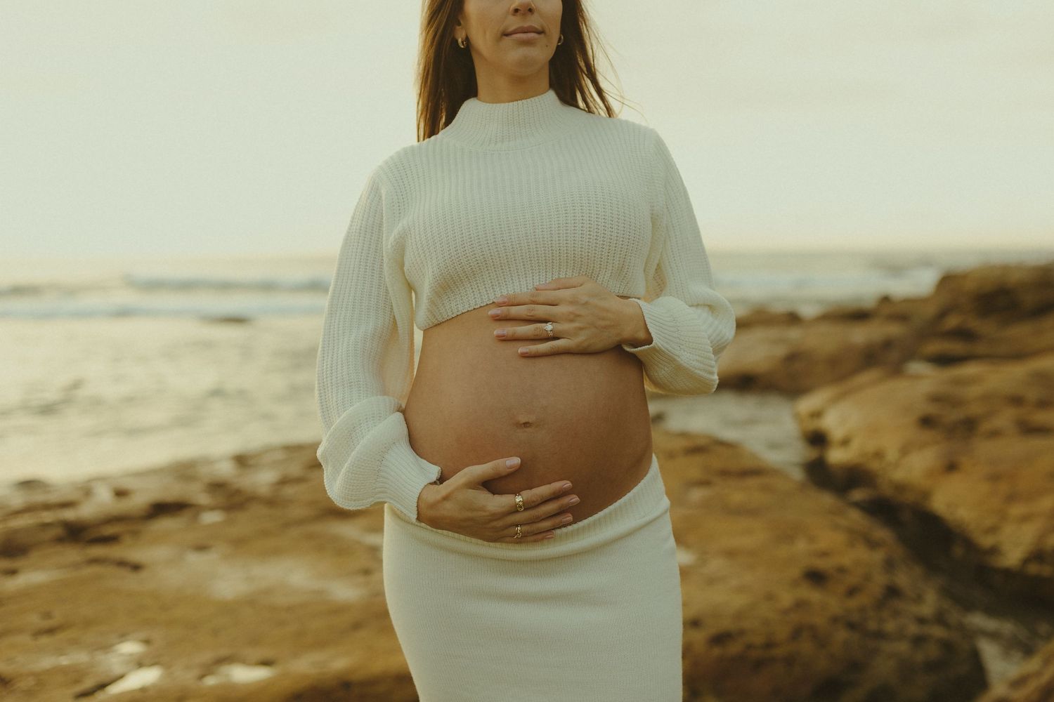 A pregnant figure in white dress holds their belly while standing on a beach at sunset.