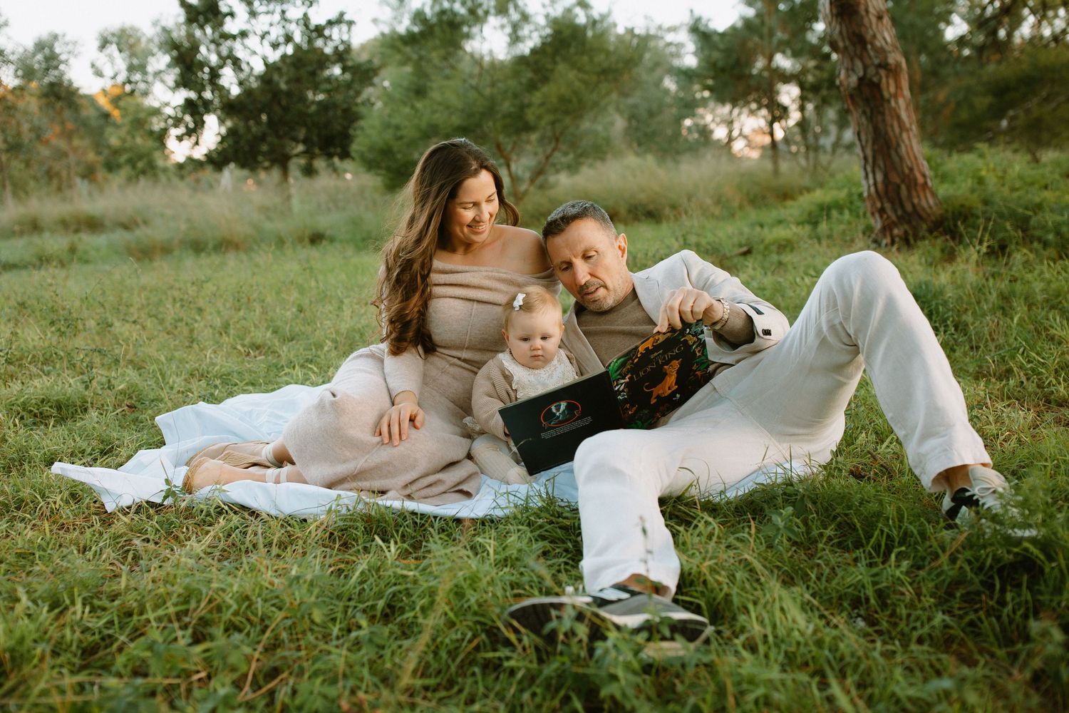 Family relaxes together on white blanket during outdoor photoshoot in grassy field.