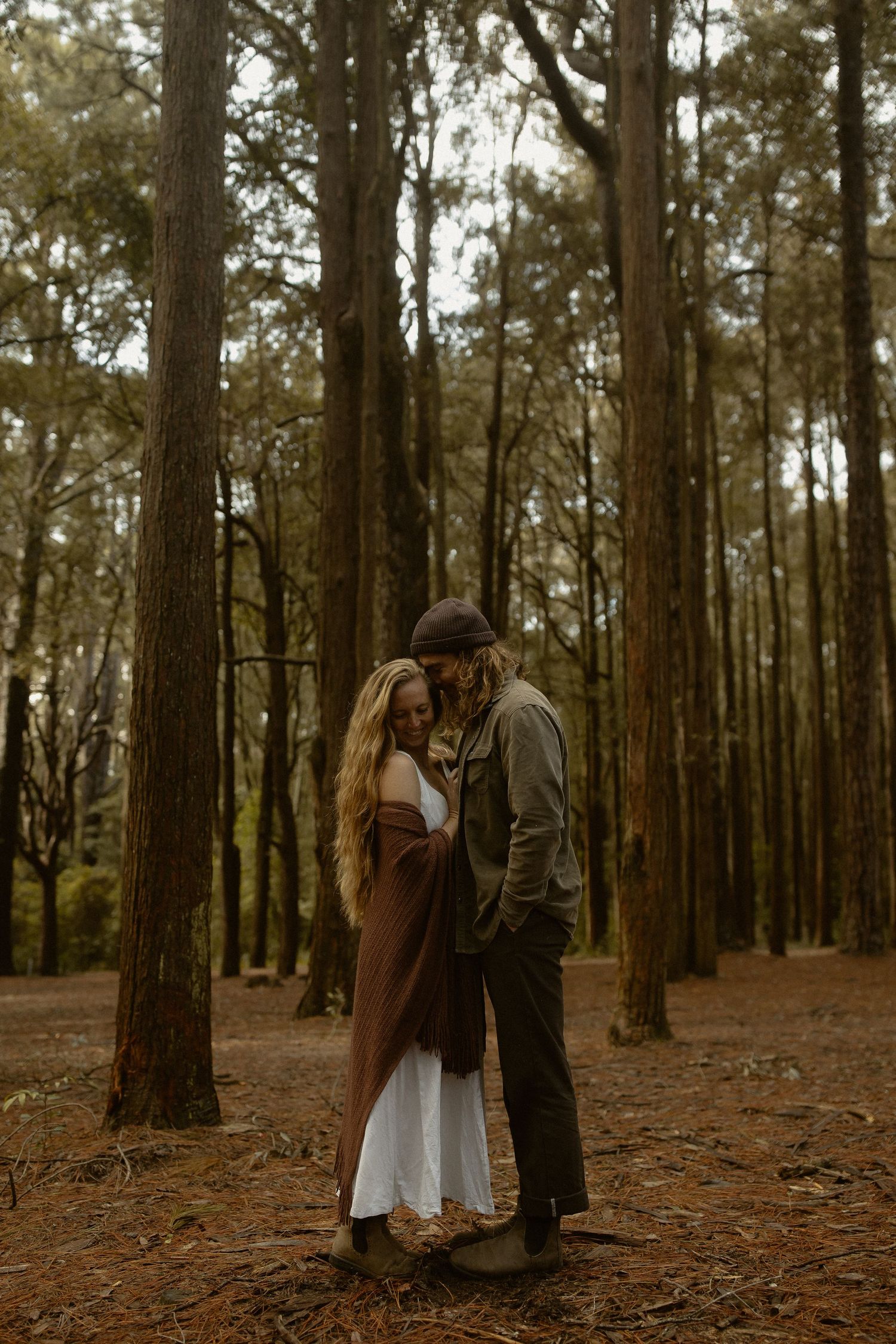 A couple shares an intimate moment among tall pine trees in a moody forest setting.