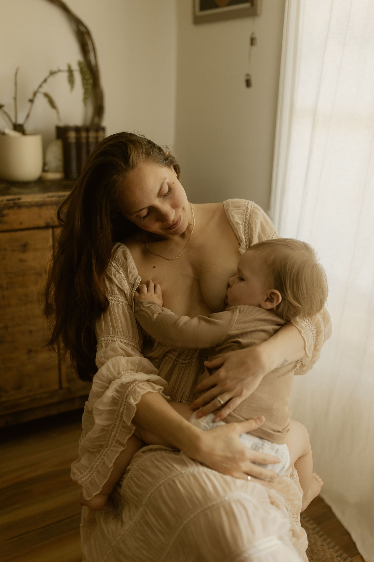 Intimate motherhood portrait in warm lighting showing nursing moment beside antique dresser.