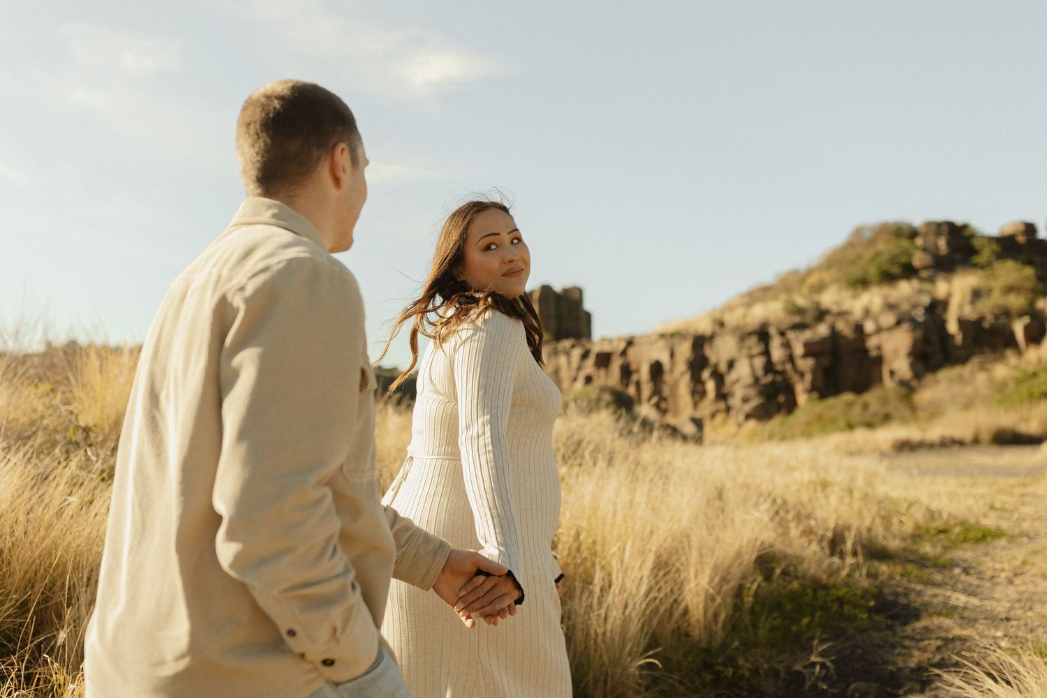 A couple share an intimate moment among tall grass with rocky cliffs in the background.