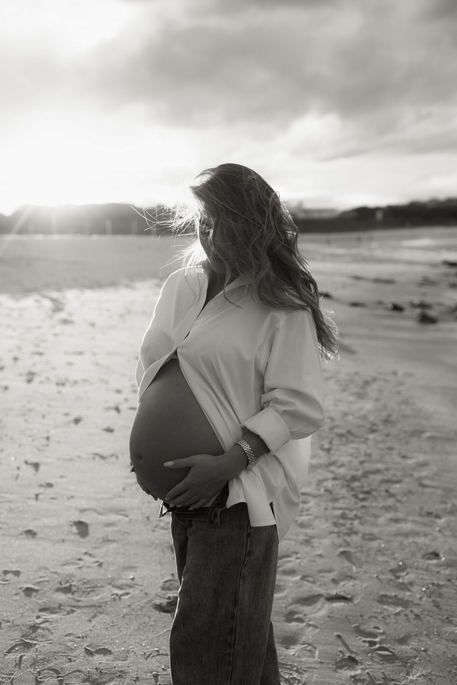 Pregnant woman in white shirt holding belly on windy beach, black and white photo.