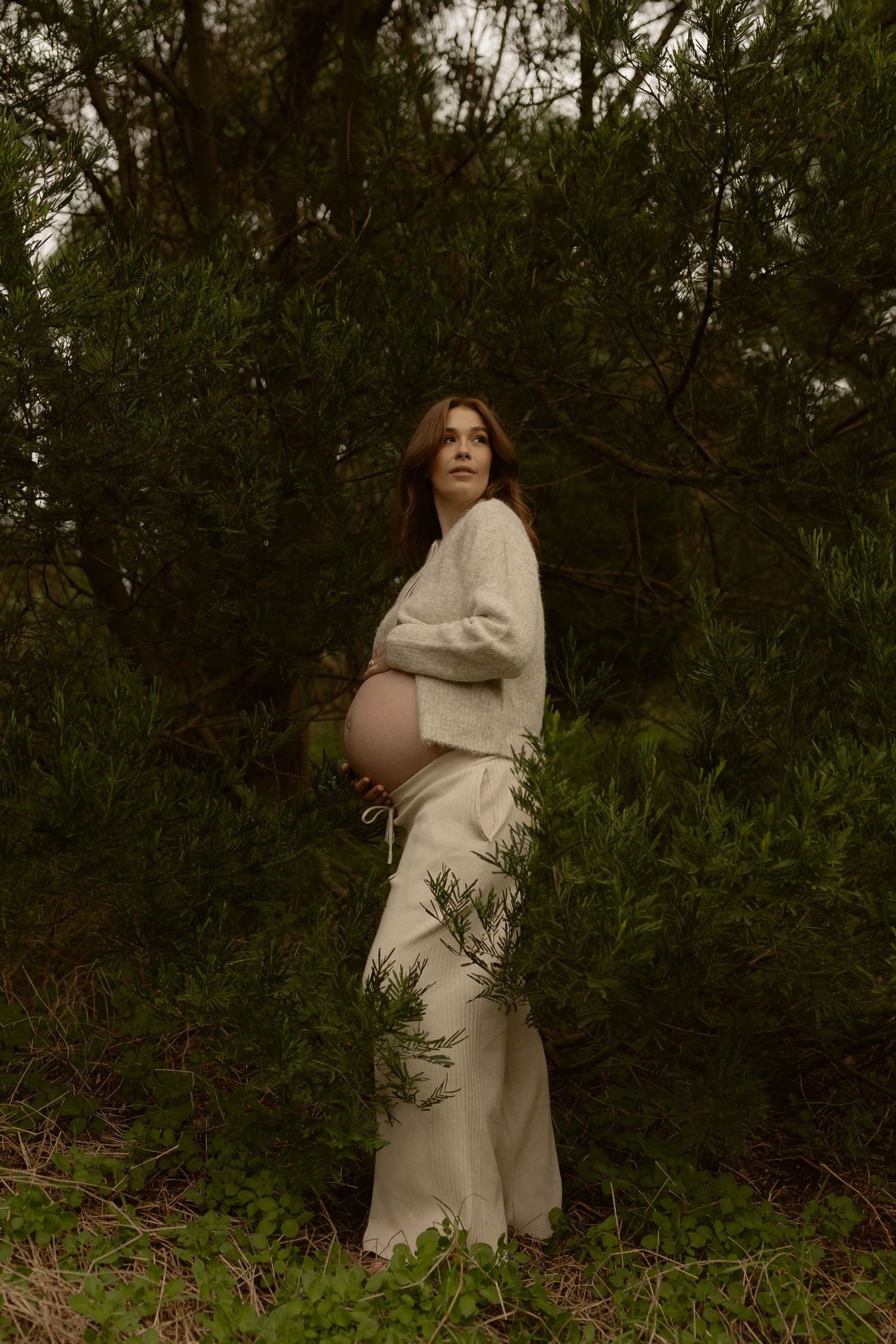 Pregnant woman in white skirt cradling bare baby bump surrounded by lush green trees outdoors.
