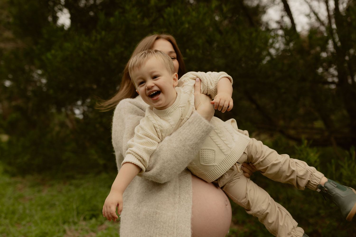 Laughing toddler being held by pregnant woman in beige sweater against lush green background.