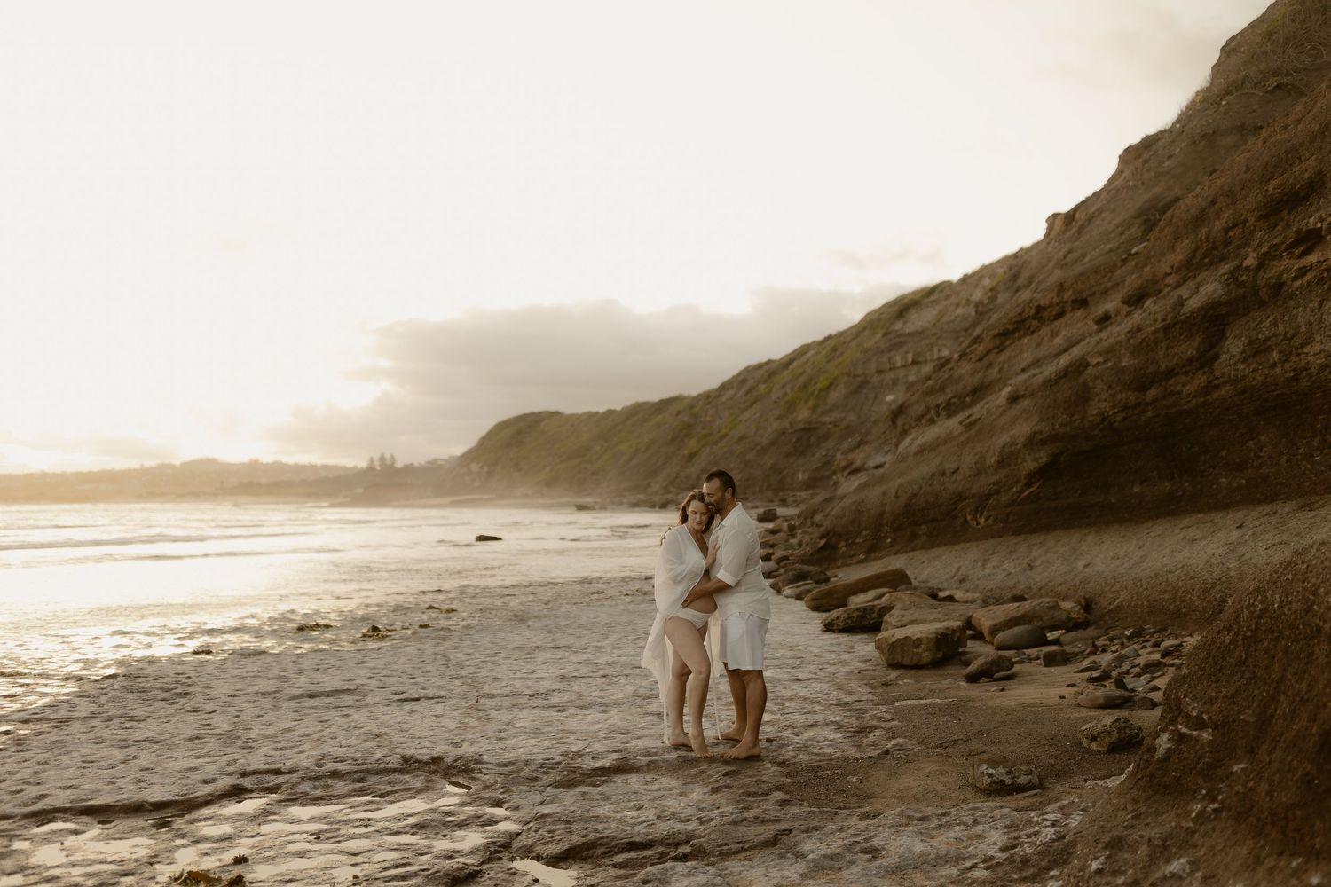 Serene beach scene with couple in white clothing walking along the sandy shore at sunset.