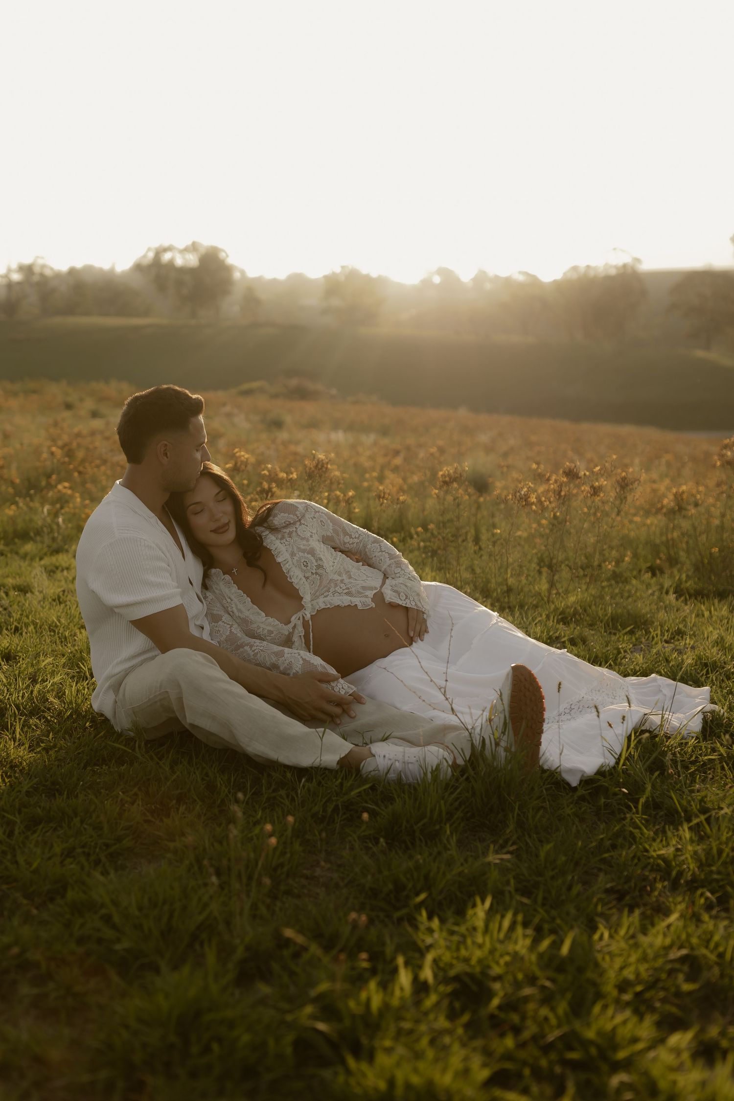 Couple lounges together in a grassy meadow at sunset wearing white clothing.