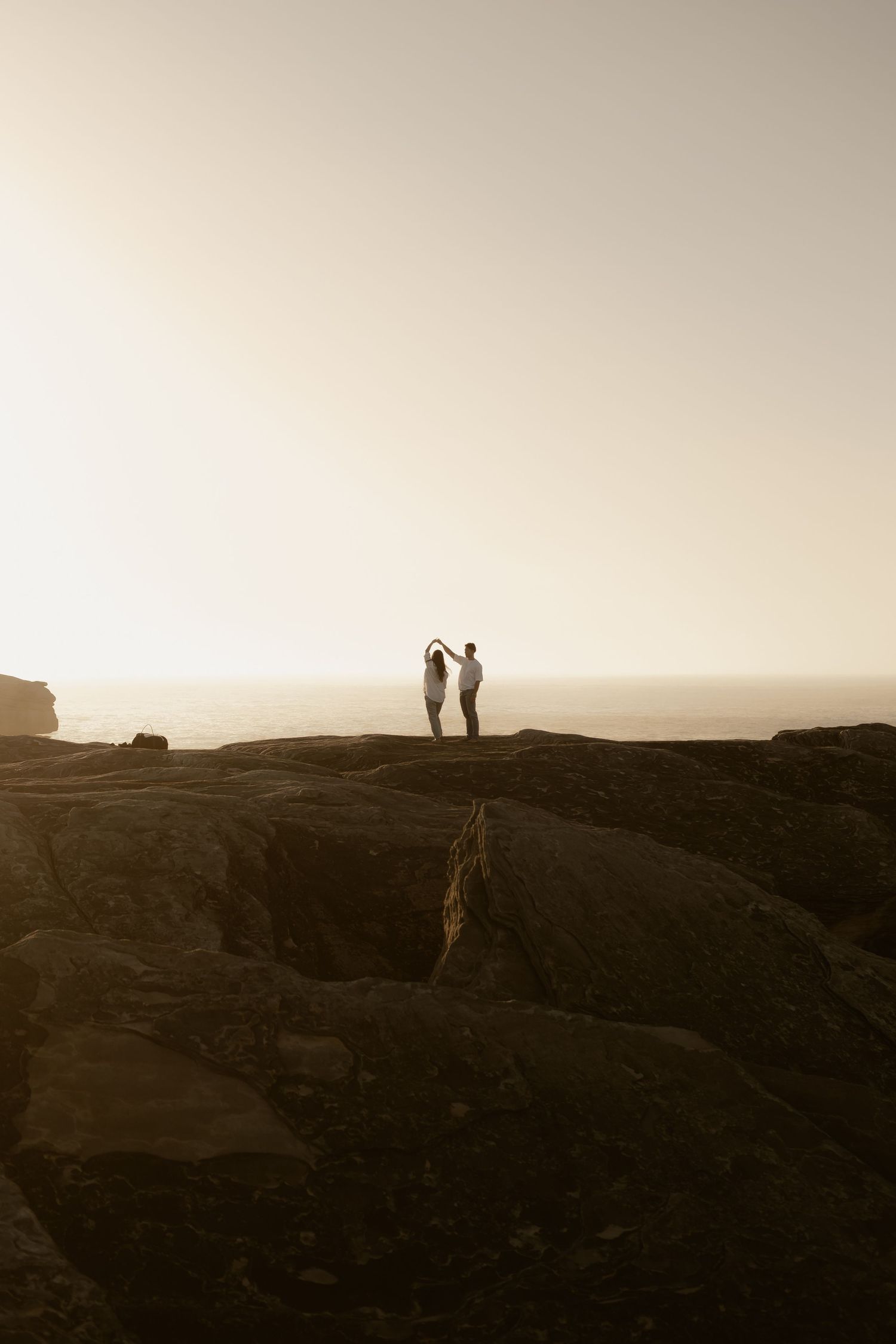 Two silhouetted figures stand together on a rocky cliff edge against a bright sunset sky.