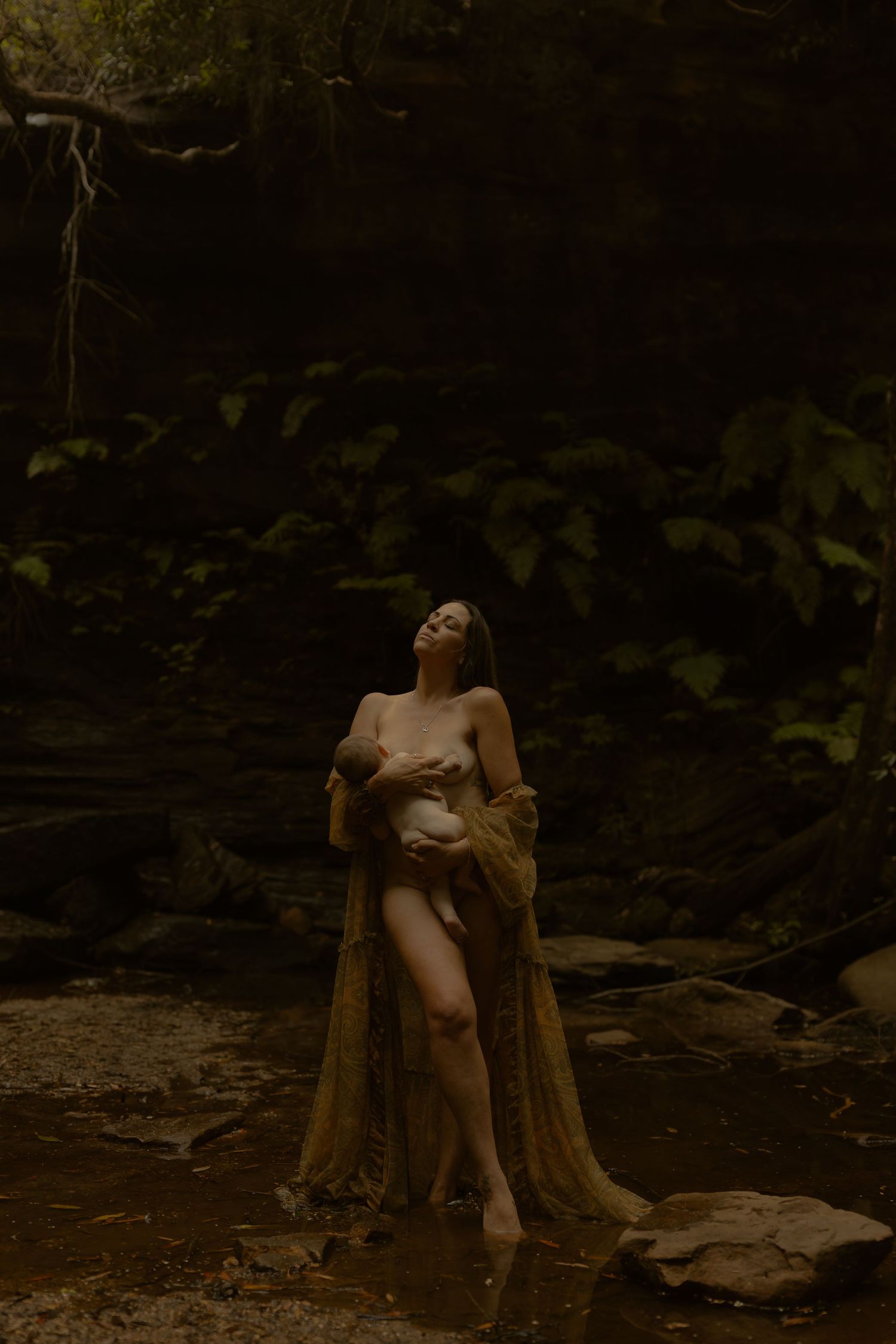 Artistic nude figure rests against a fallen tree trunk in a dark moody forest setting.
