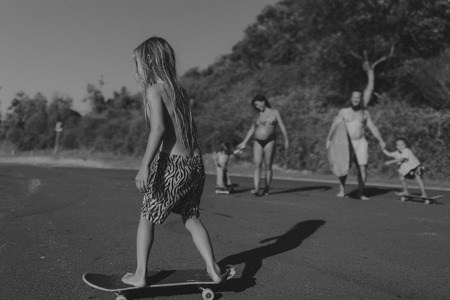 People skateboarding on a coastal road with long shadows cast in black and white.