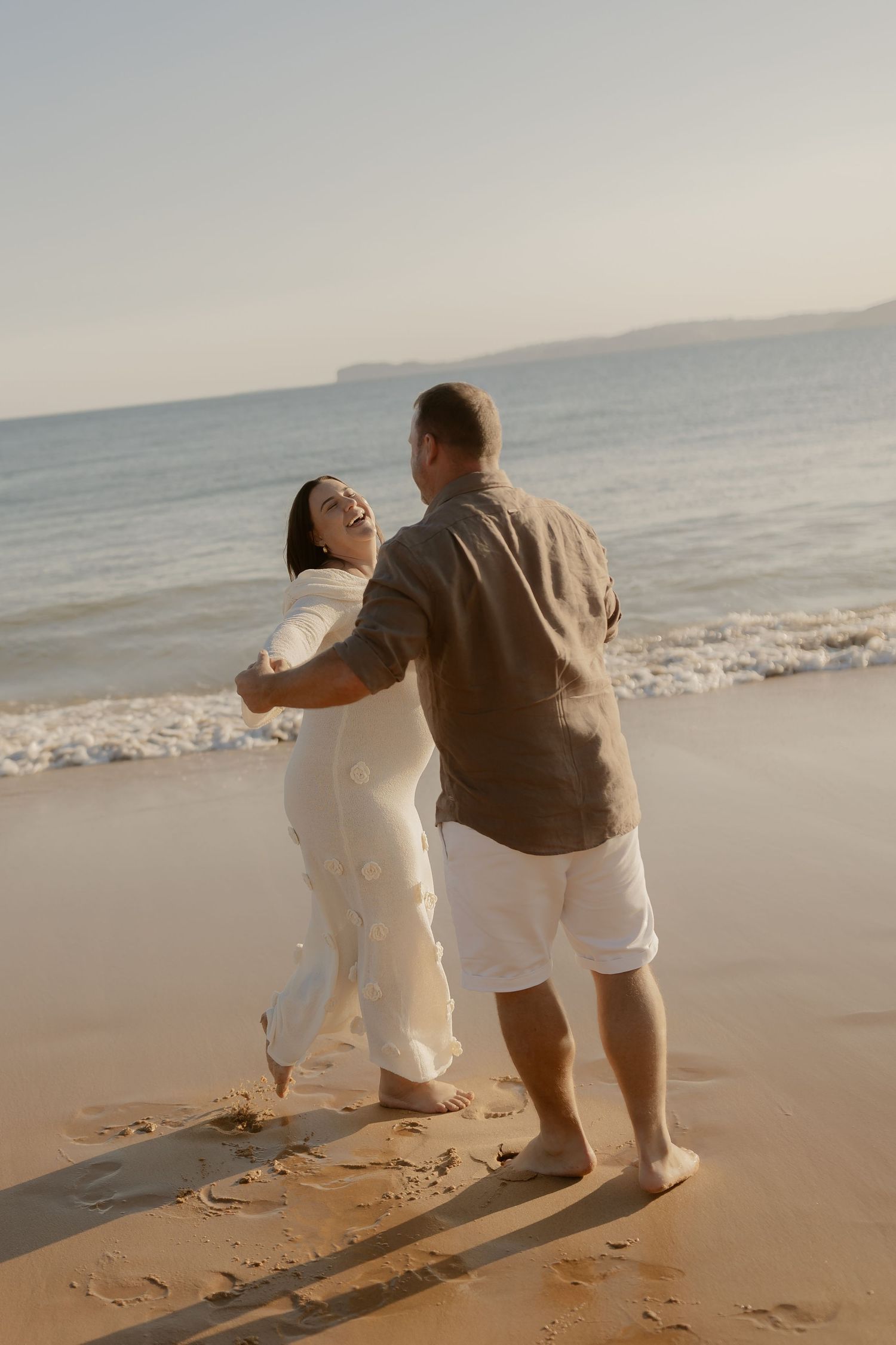 A couple shares a romantic moment dancing together on a sunlit beach with ocean waves in the background.