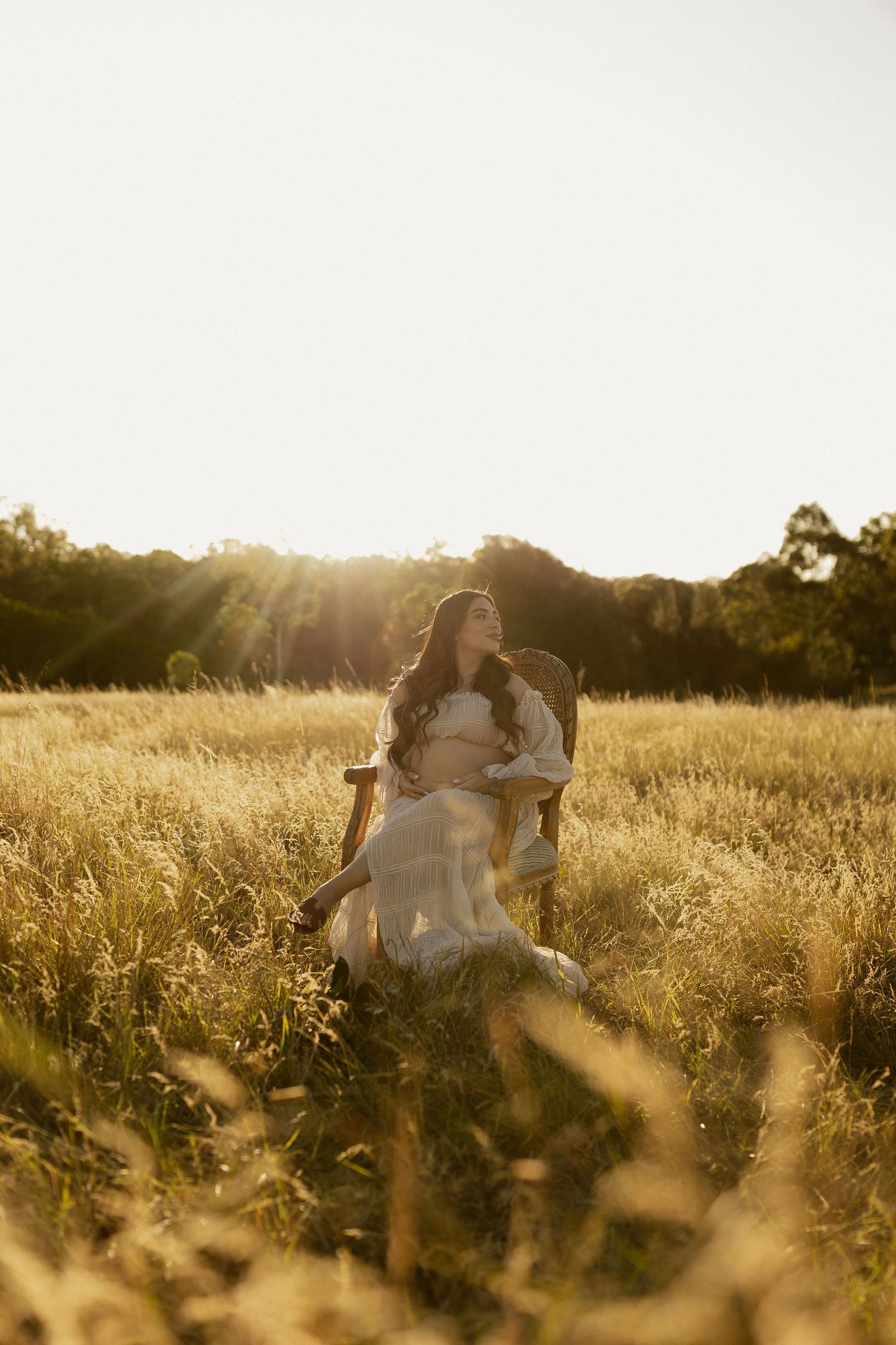 A sunlit maternity portrait captures a tender moment in a rustic field with golden hour lighting.