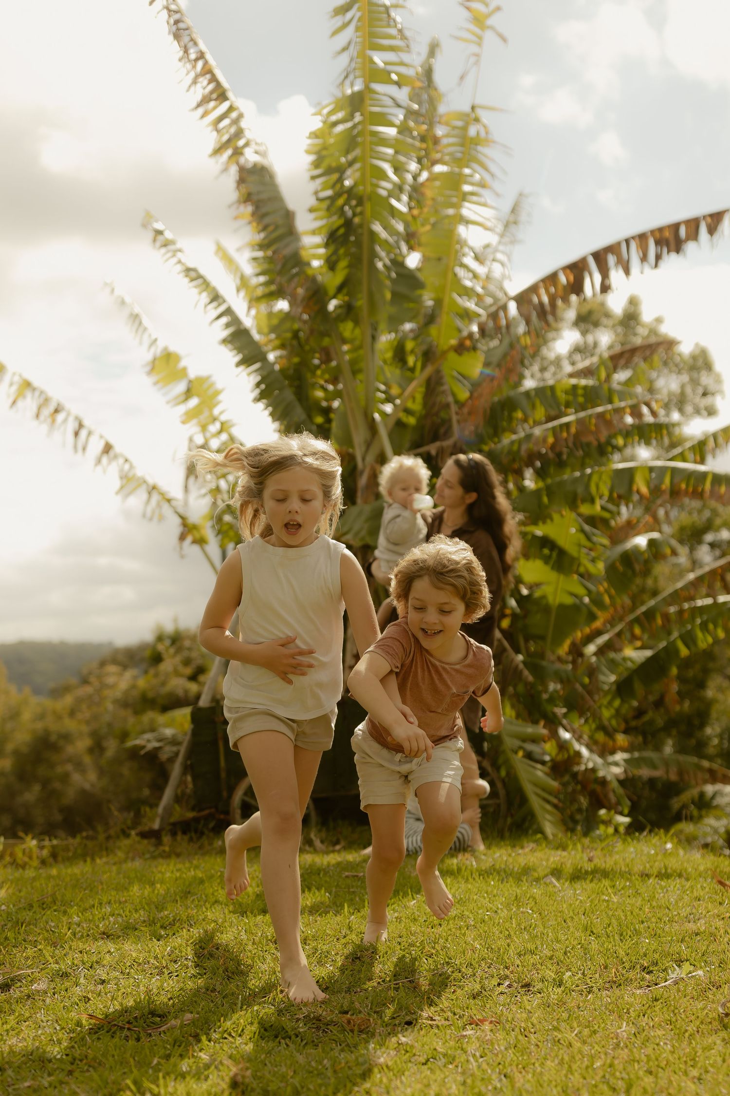 Two barefoot children run joyfully on green grass with a woman and toddler near tropical palm trees outdoors.
