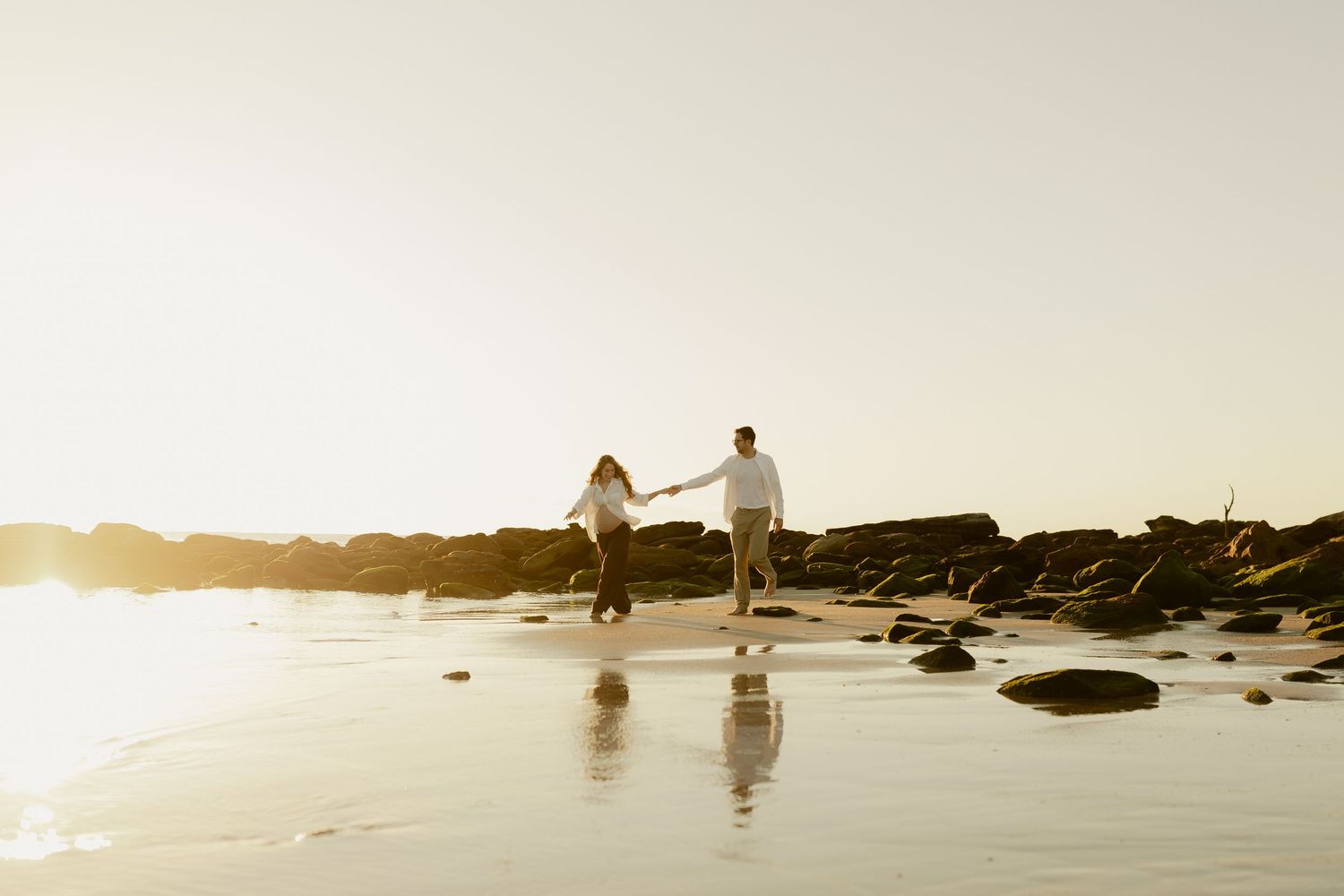 Couple holding hands walking along sandy beach with moss-covered rocks at golden sunset.